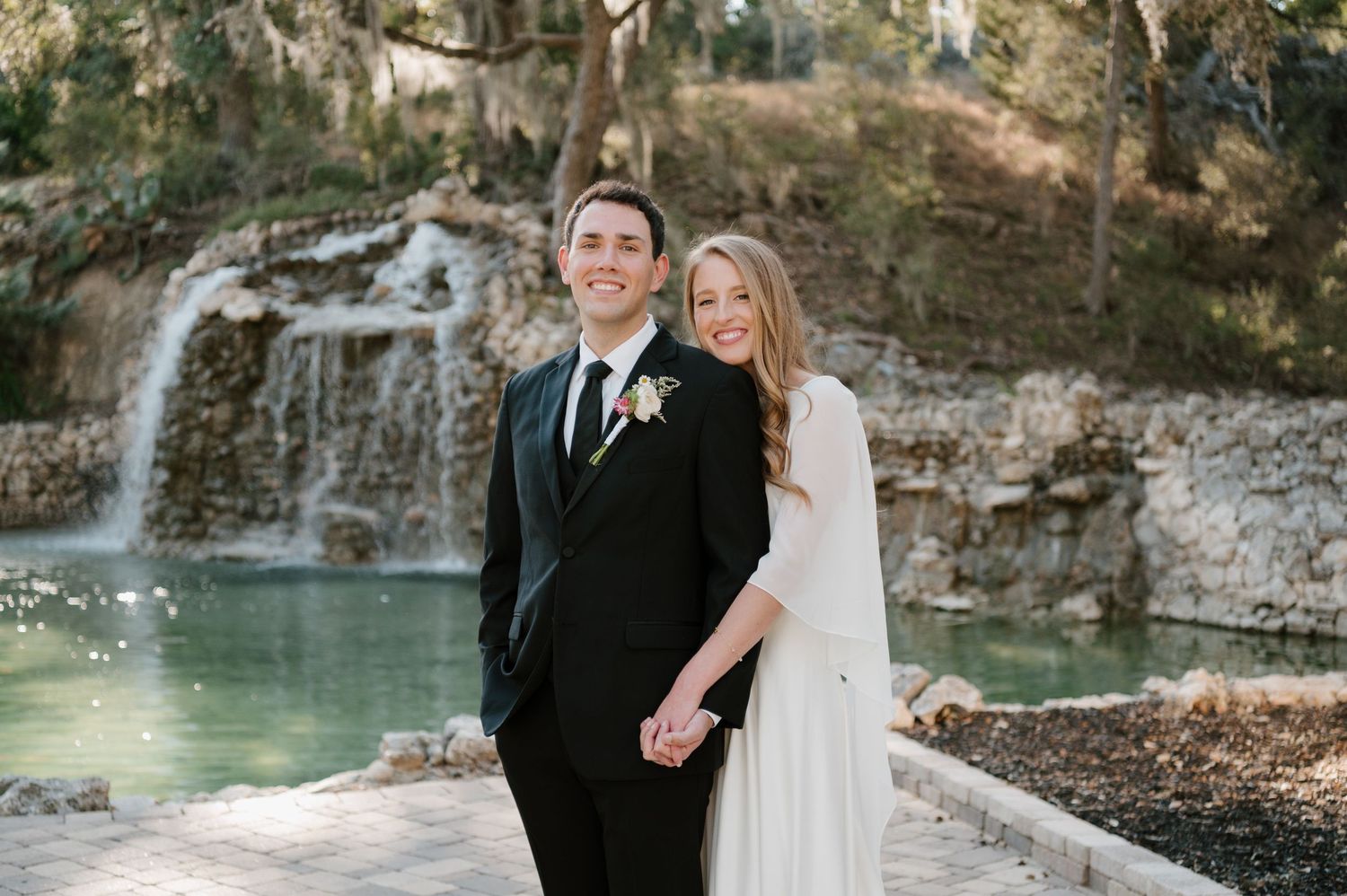 A couple poses near a serene waterfall and turquoise pool surrounded by rocky cliffs in a romantic outdoor setting.