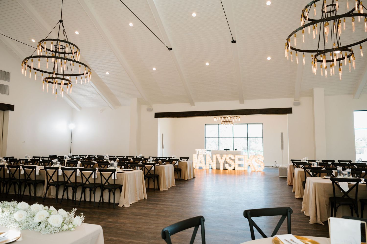 Modern white wedding reception venue with elegant circular chandeliers and cross-back chairs arranged in rows.