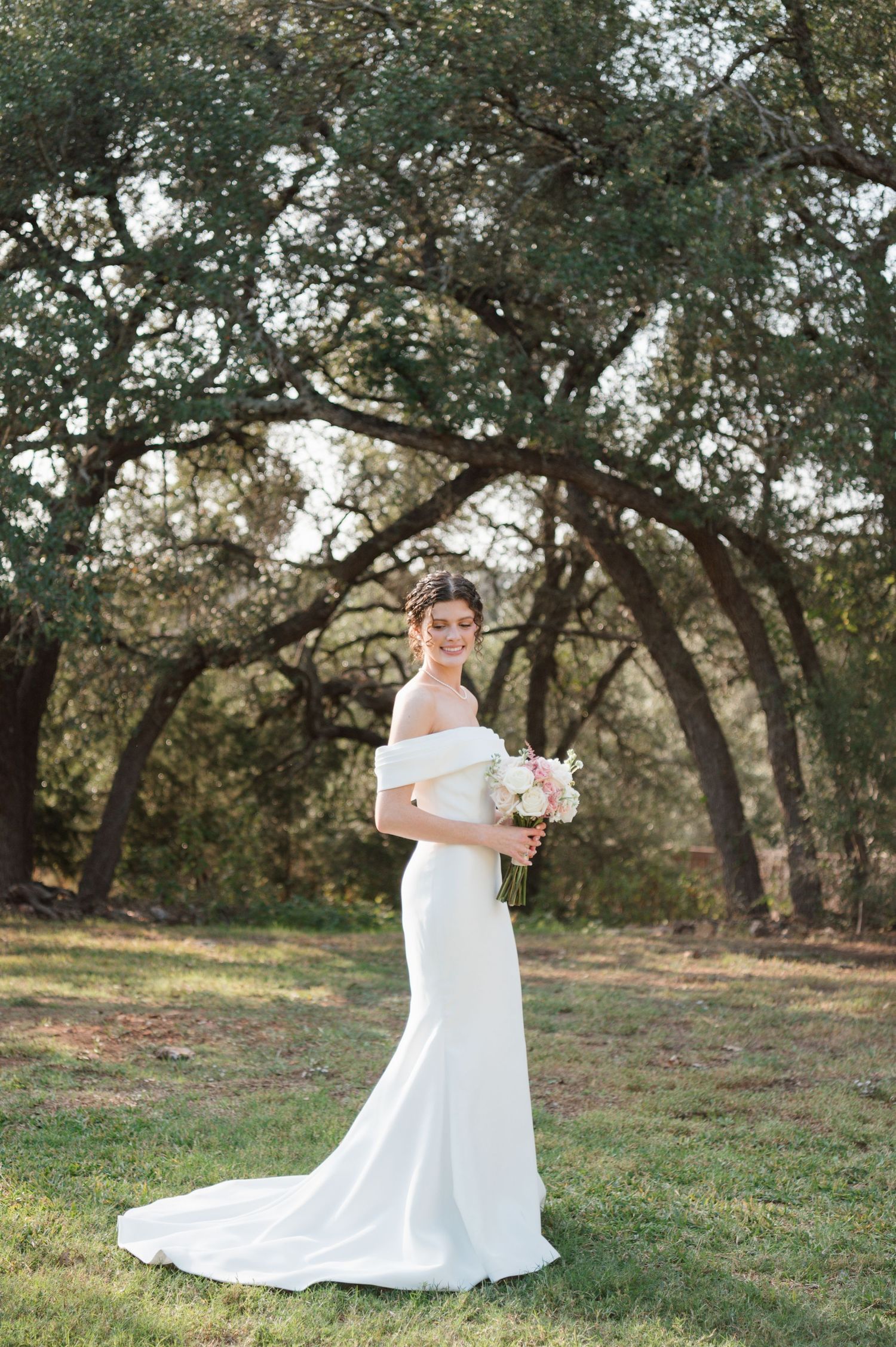 Elegant bride in off-shoulder white gown poses outdoors under oak trees holding pink and white bouquet.