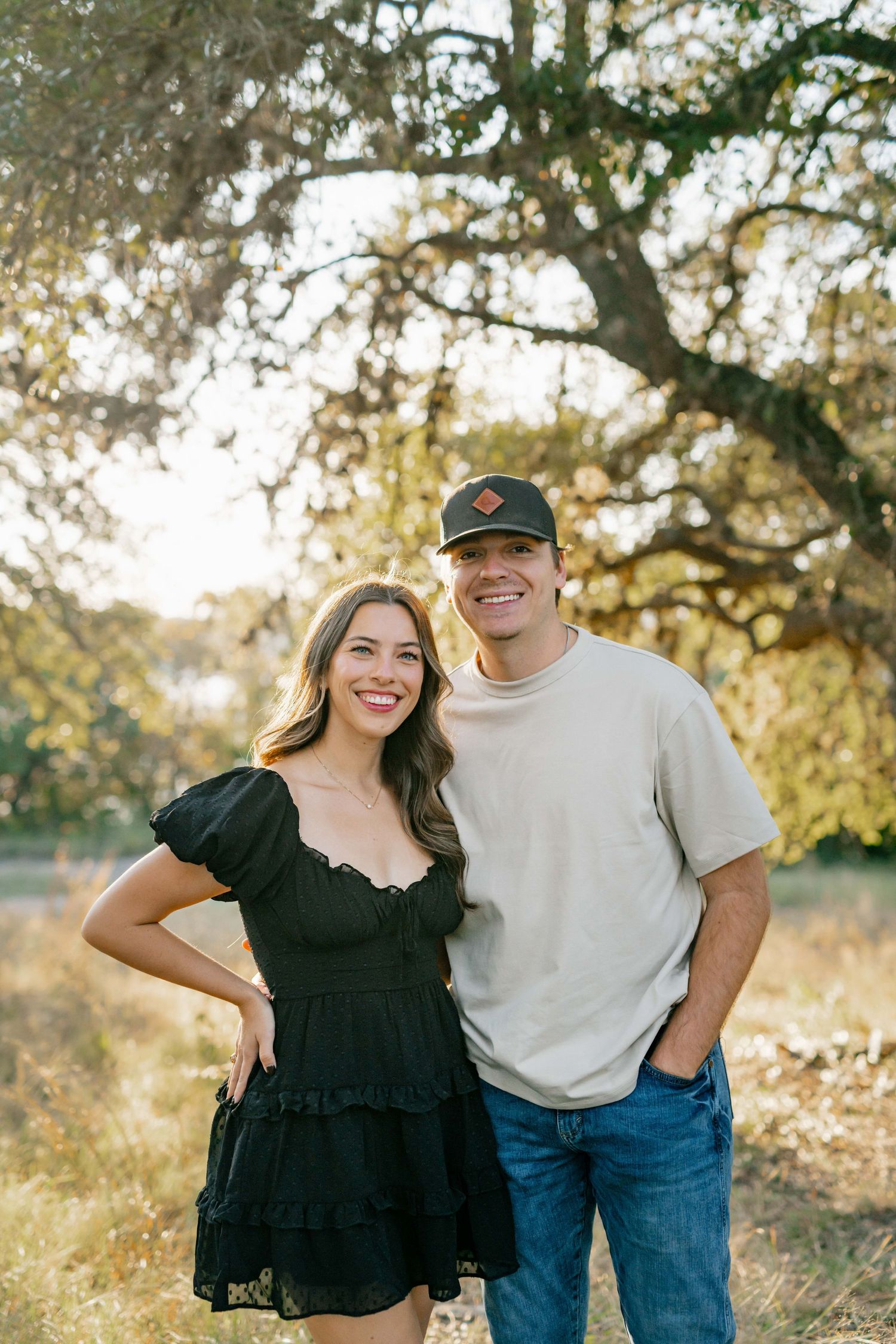 A couple stands together outdoors with golden sunlight filtering through trees behind them.