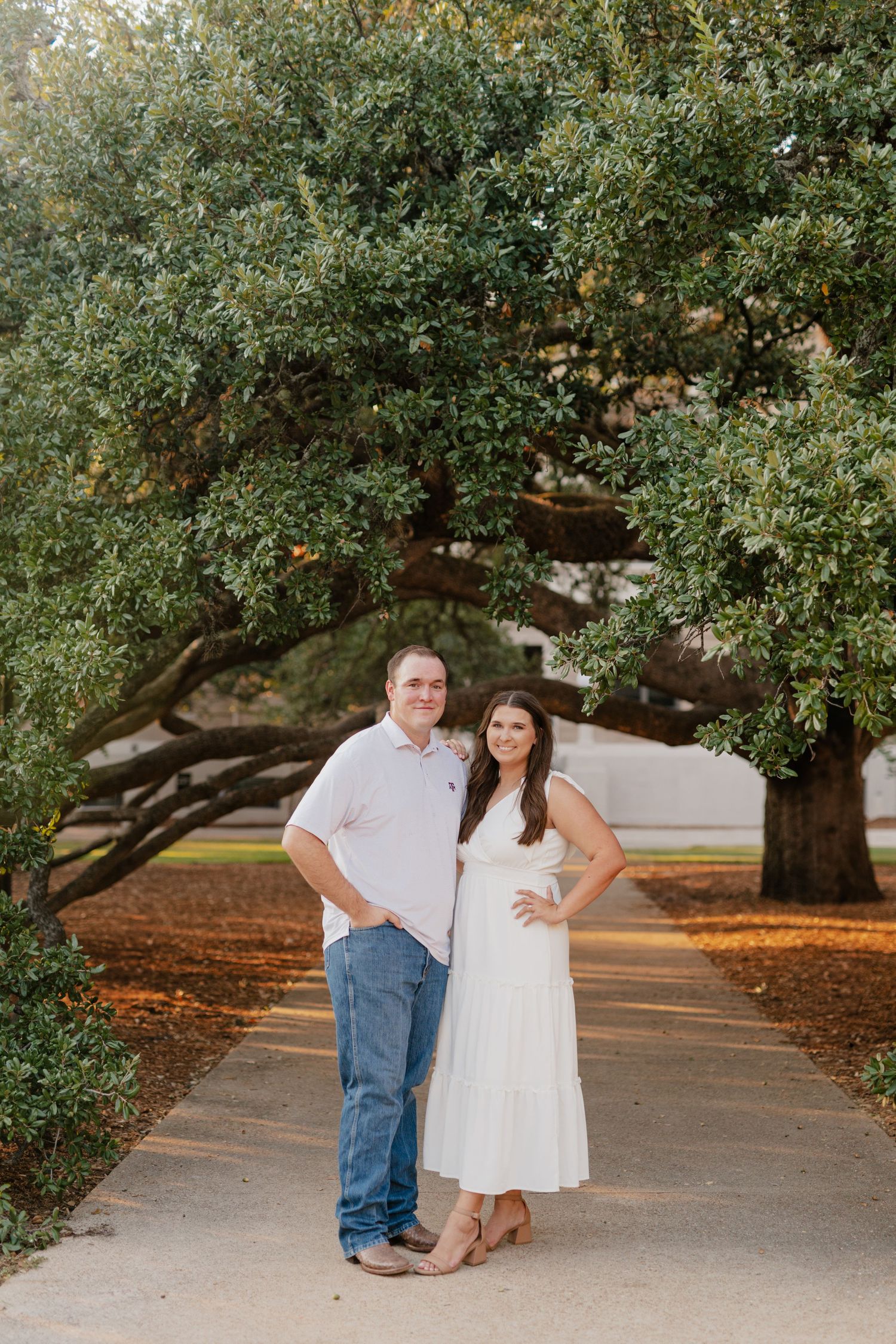 A couple poses together under a sprawling oak tree on a paved pathway.