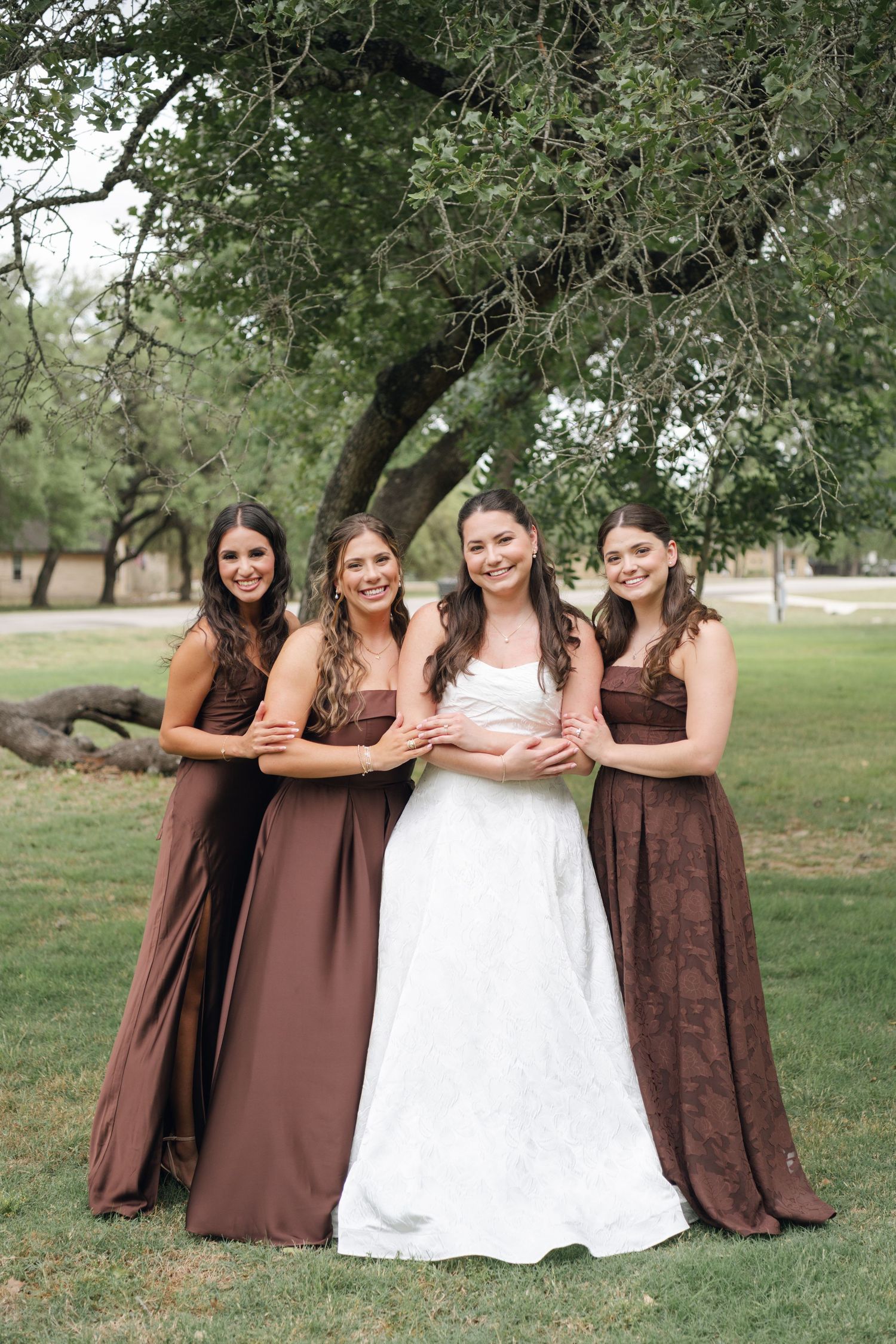 Bride in white gown poses with three bridesmaids in brown dresses under a large tree outdoors.