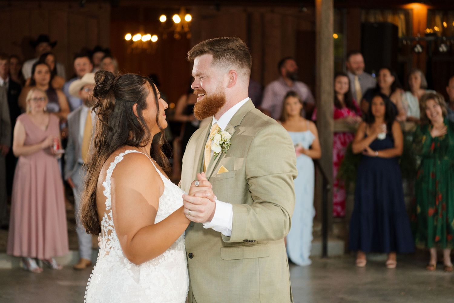 Bride and groom share their first dance surrounded by guests in a warmly lit rustic barn reception.