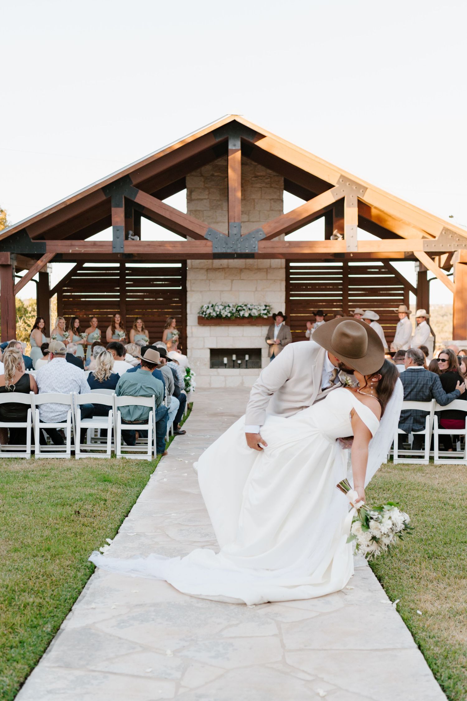 A romantic outdoor wedding ceremony takes place under a rustic wooden pavilion with white chairs and guests gathered.