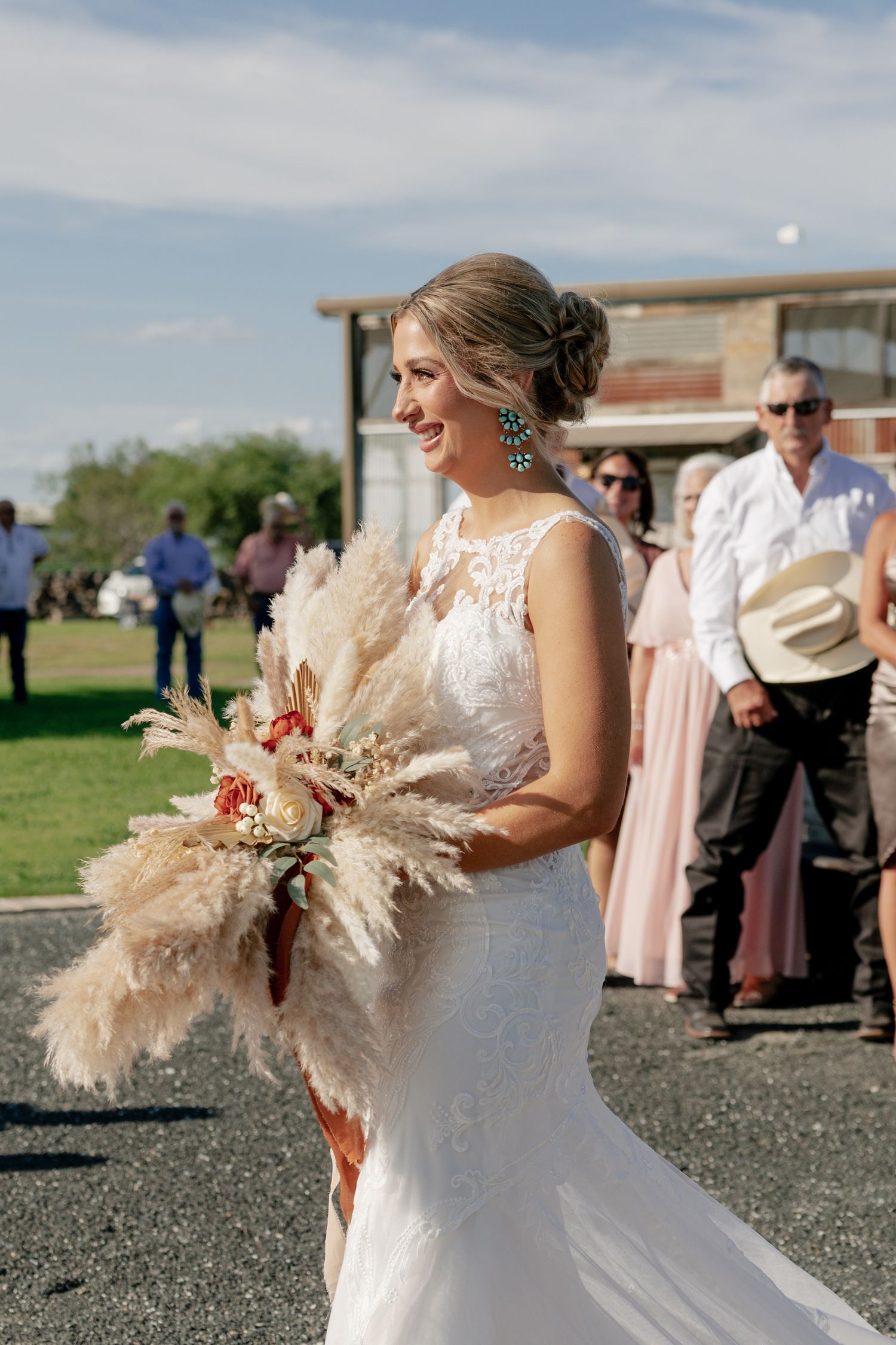 A bride in a lace gown holding a pampas grass bouquet at an outdoor ceremony.