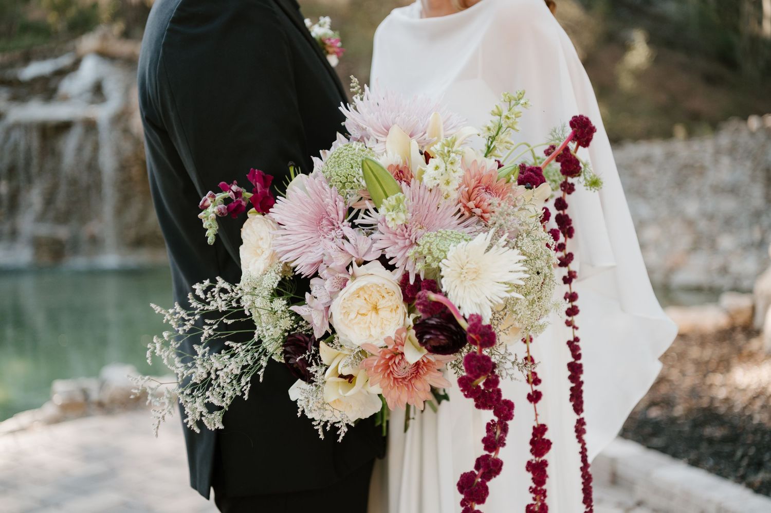 Romantic wedding bouquet featuring pink, burgundy and white flowers with cascading amaranthus against a lakeside backdrop.