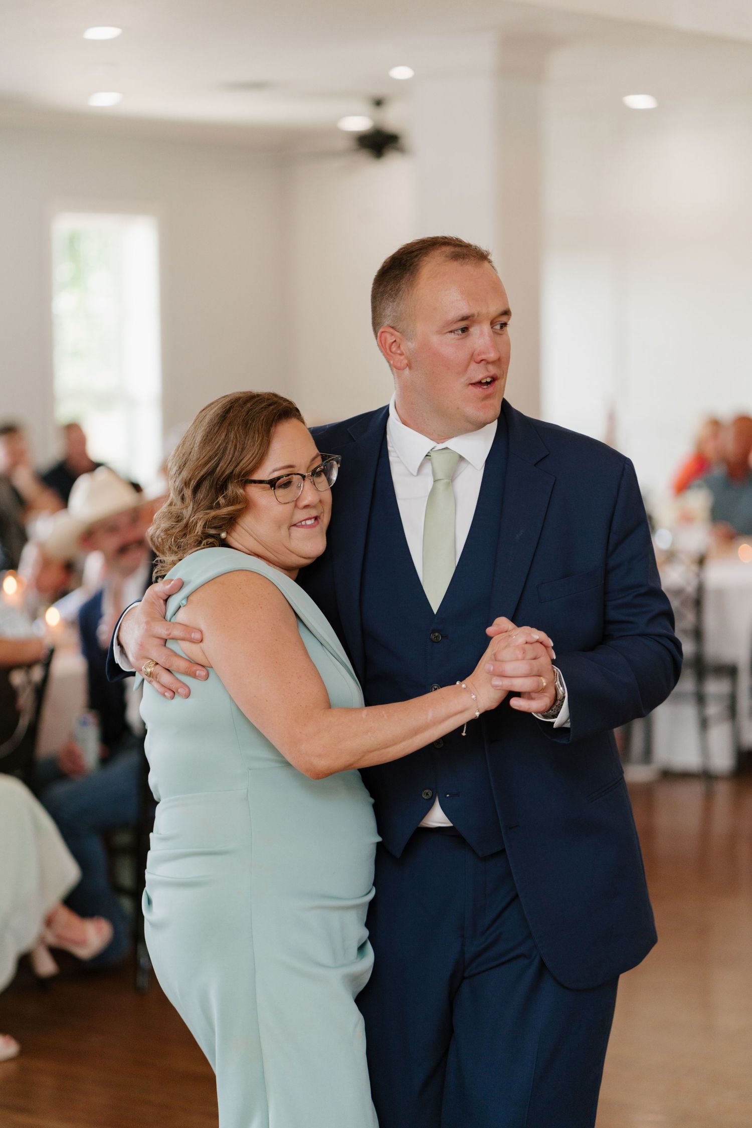 Guests in formal attire dance together on a bright wooden dance floor at a wedding reception.