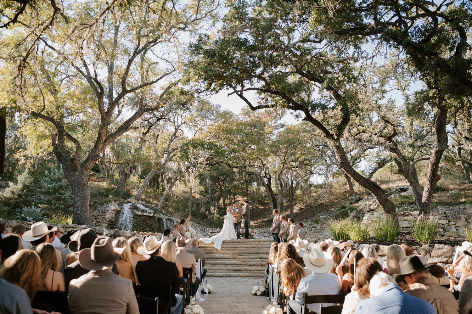 Wedding ceremony guests seated outdoors under trees with natural lighting.