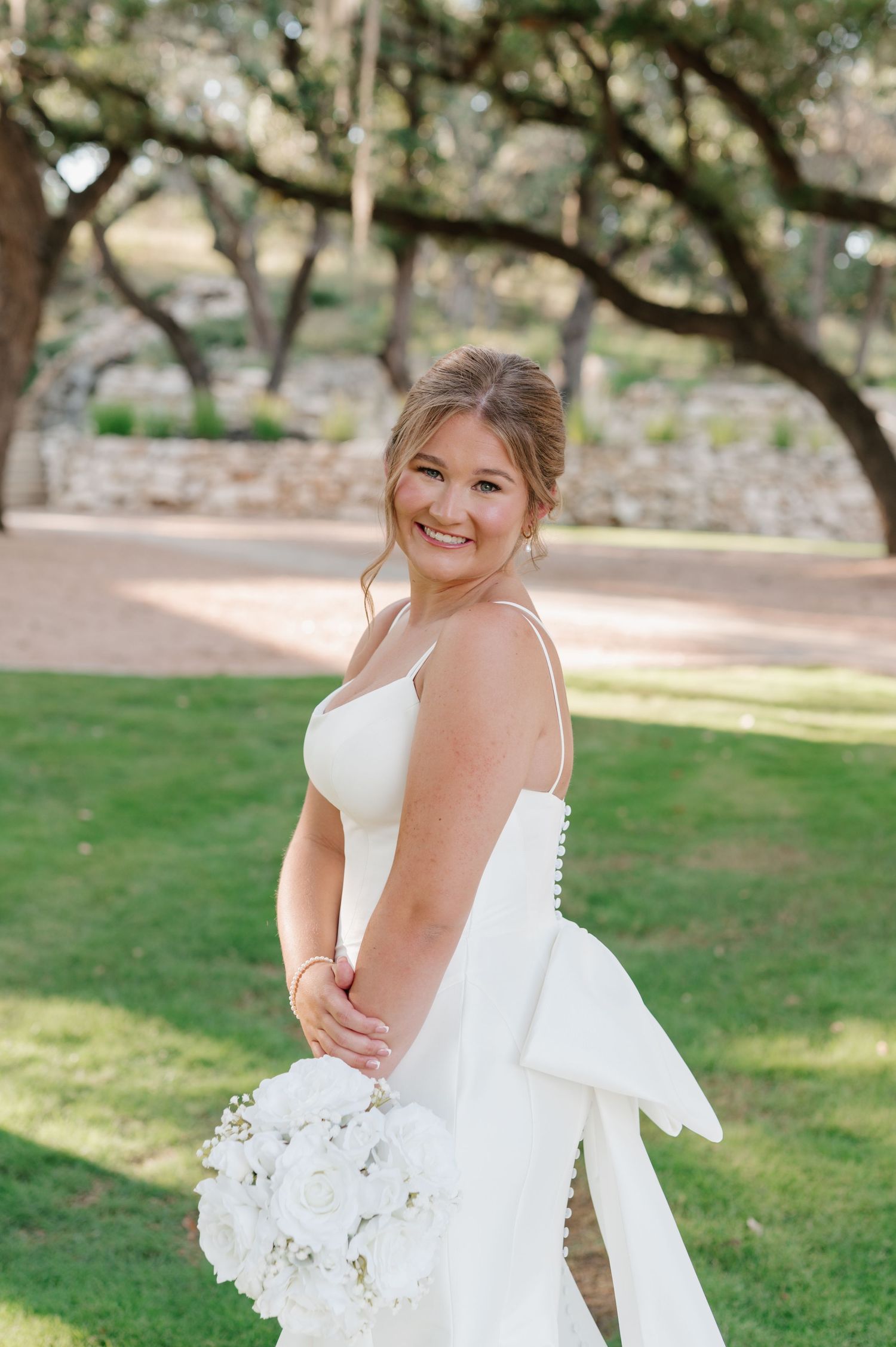 Bride in a classic white wedding dress poses outdoors under oak trees with a white bouquet.