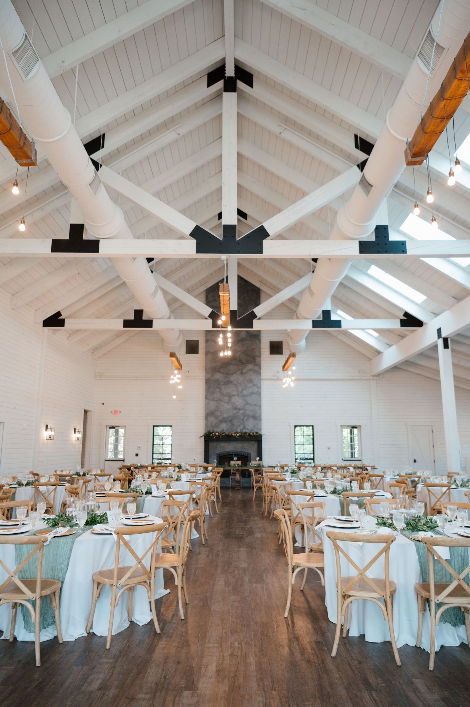 Modern white barn reception venue with vaulted ceilings, exposed beams, and cross-back chairs set for a wedding celebration.