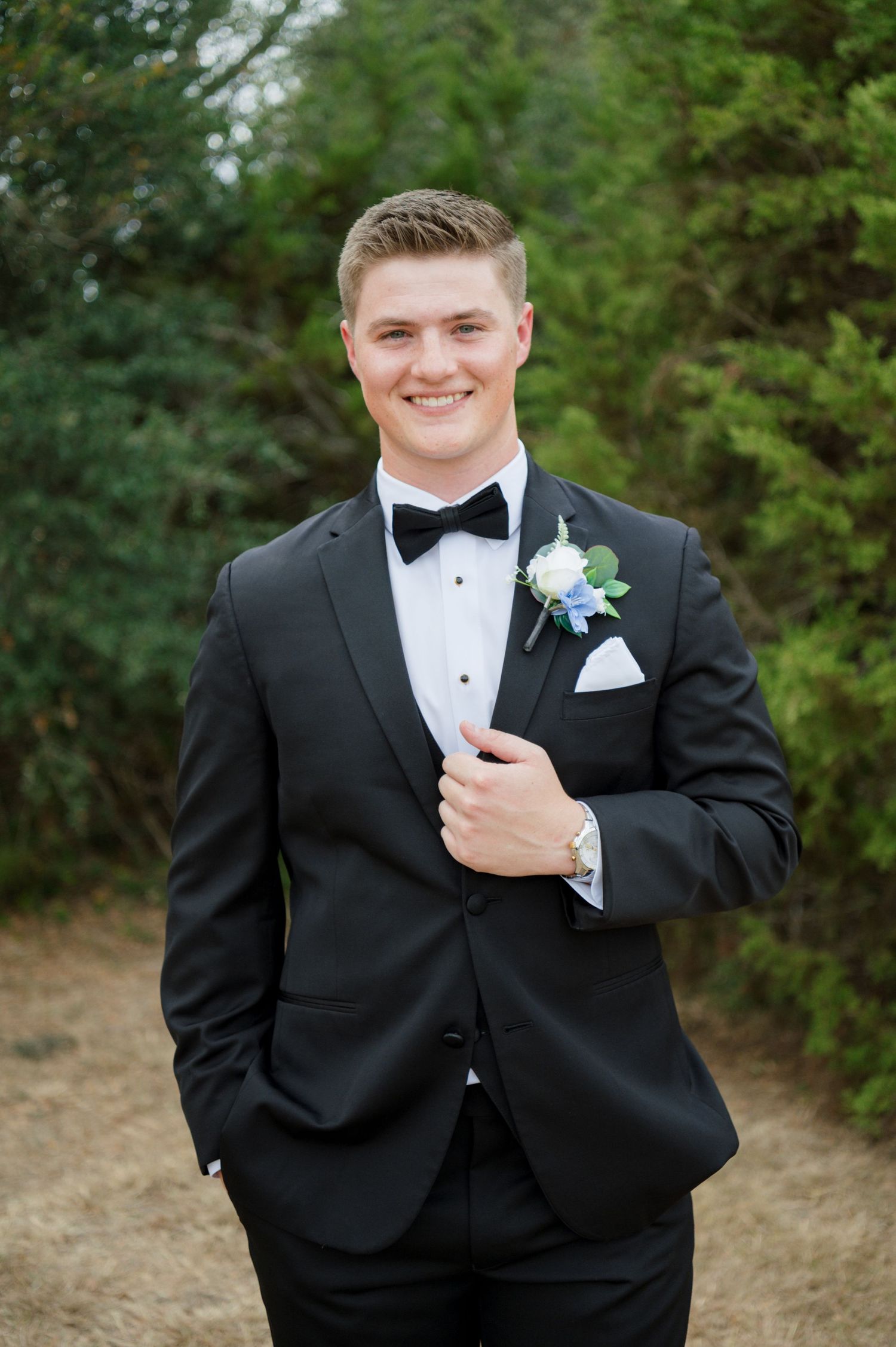 A man in a tuxedo with a boutonniere on his lapel, standing outdoors in a wooded area and smiling.