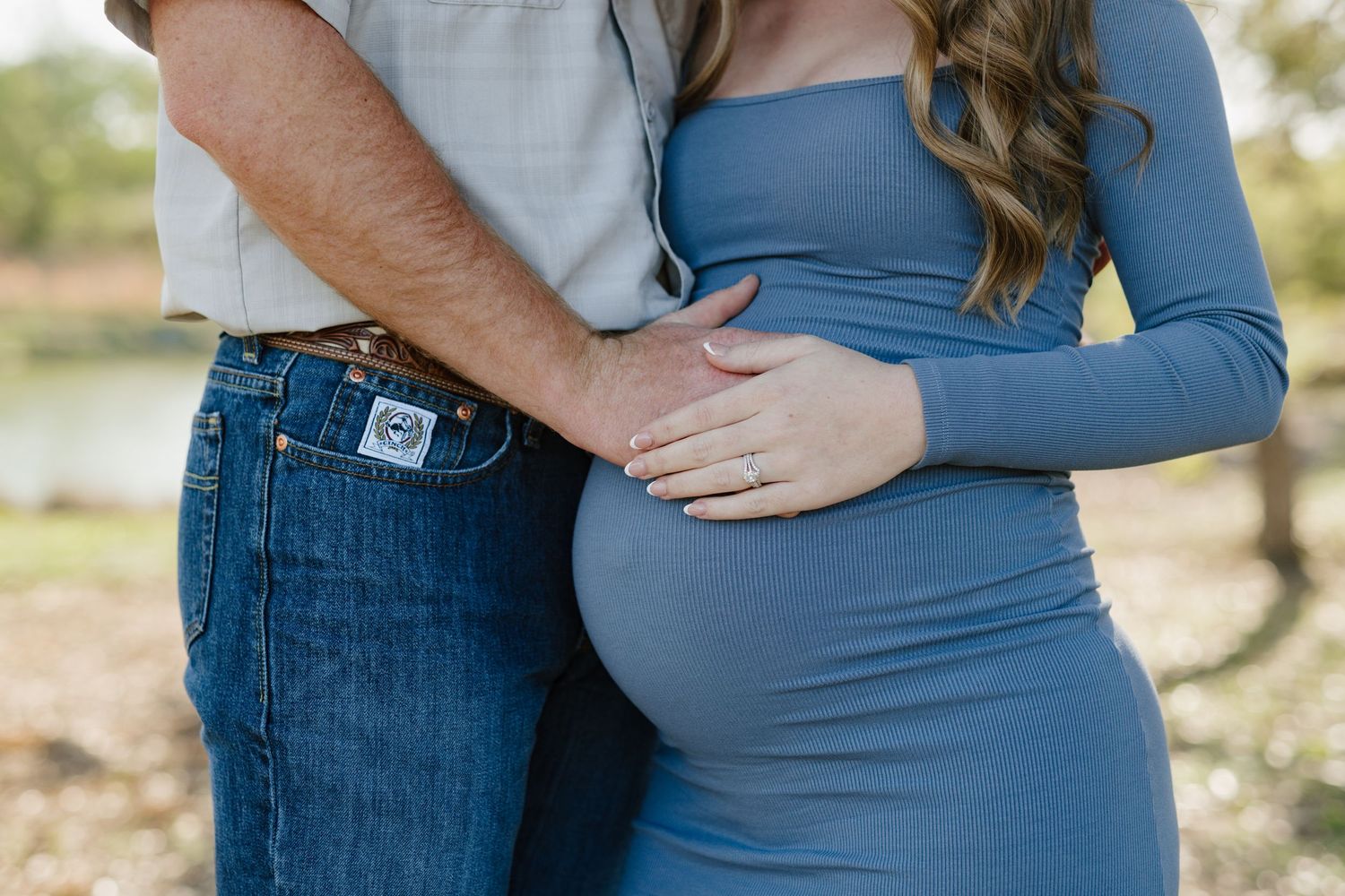 Close up of a couple's hands resting on a pregnant belly in a blue dress during an outdoor photoshoot.
