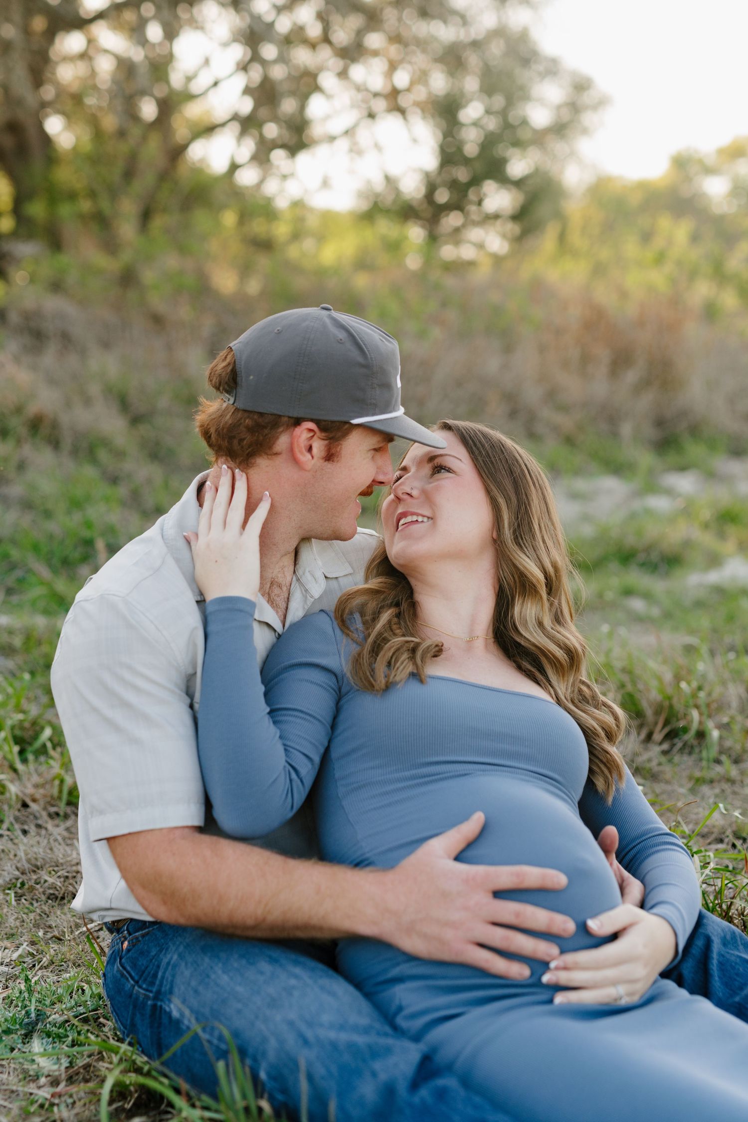 Sweet maternity photo outdoors showing couple in casual attire embracing on grass with soft natural lighting.