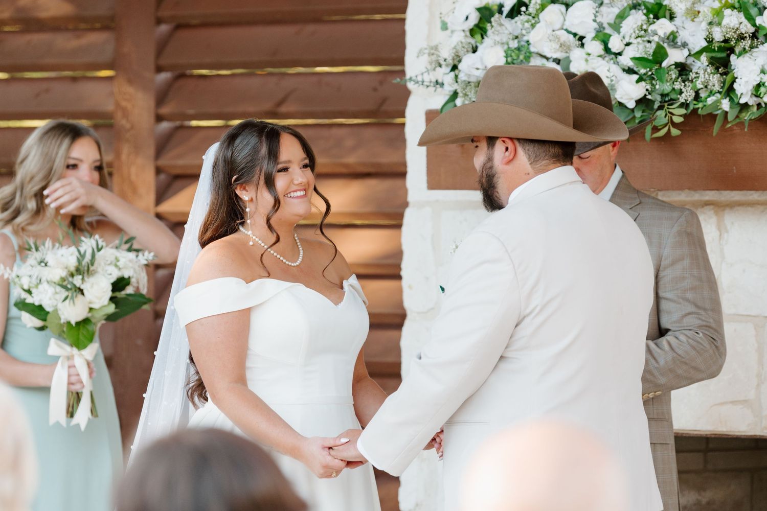 A couple participates in an intimate outdoor wedding ceremony under a rustic wooden gazebo decorated with white flowers.