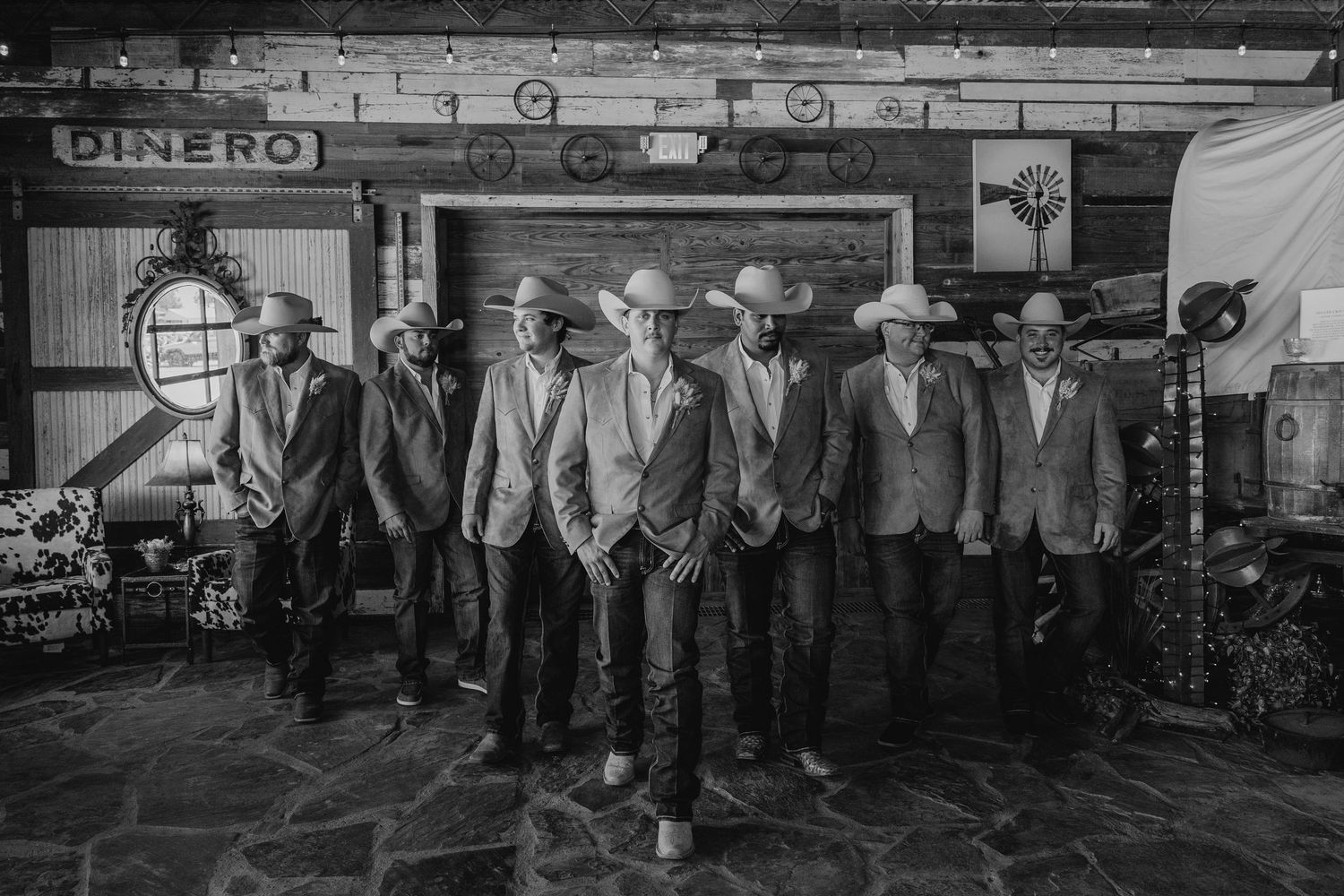 Row of groomsmen in matching suits at rustic venue in black and white photograph.