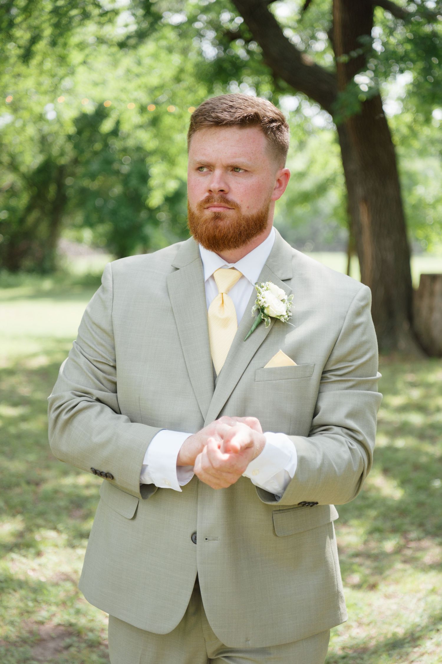 Groom in light gray suit with yellow tie and boutonniere stands outdoors among green trees on wedding day.