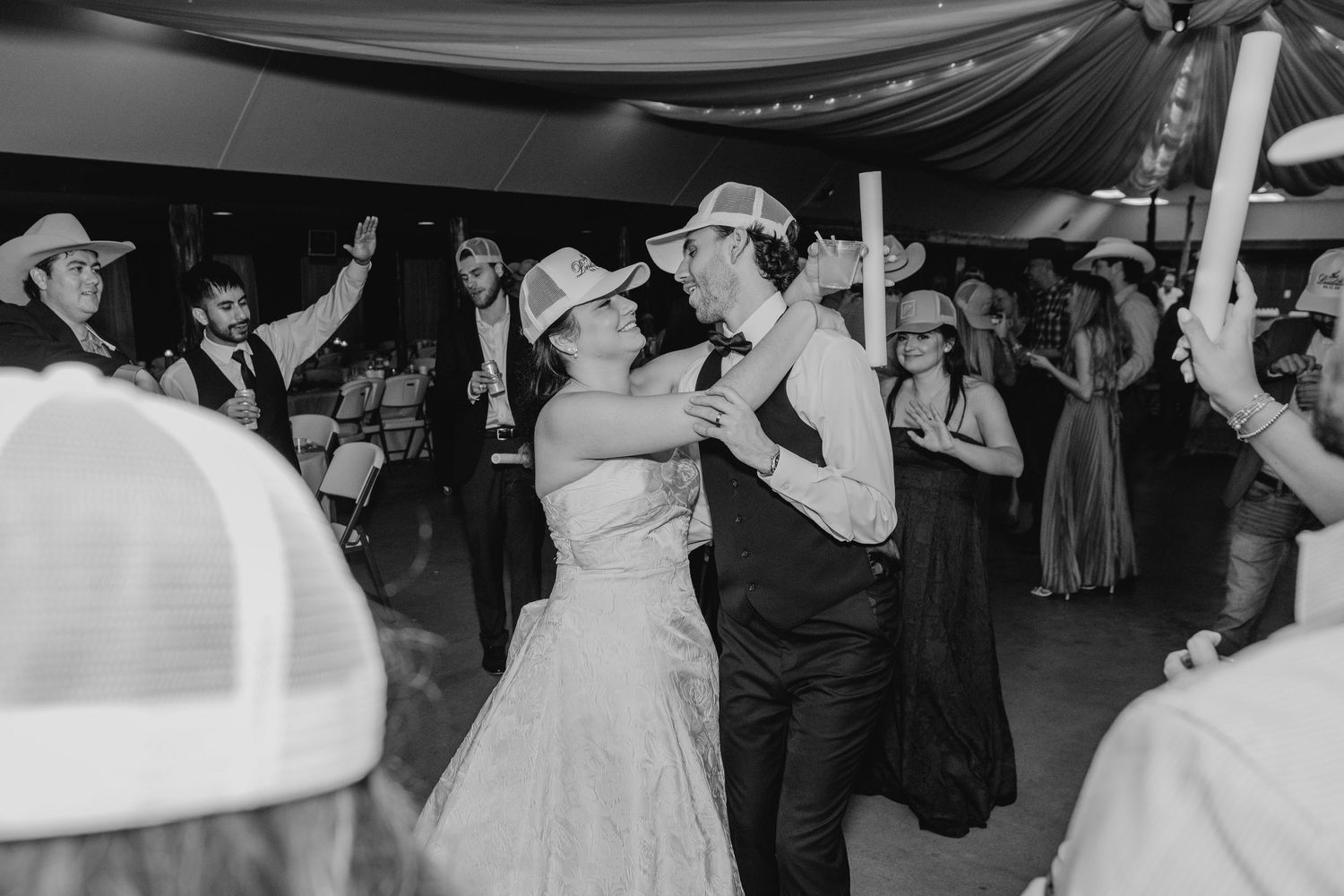 Bride and groom share romantic dance in black and white photo while guests celebrate at wedding reception.