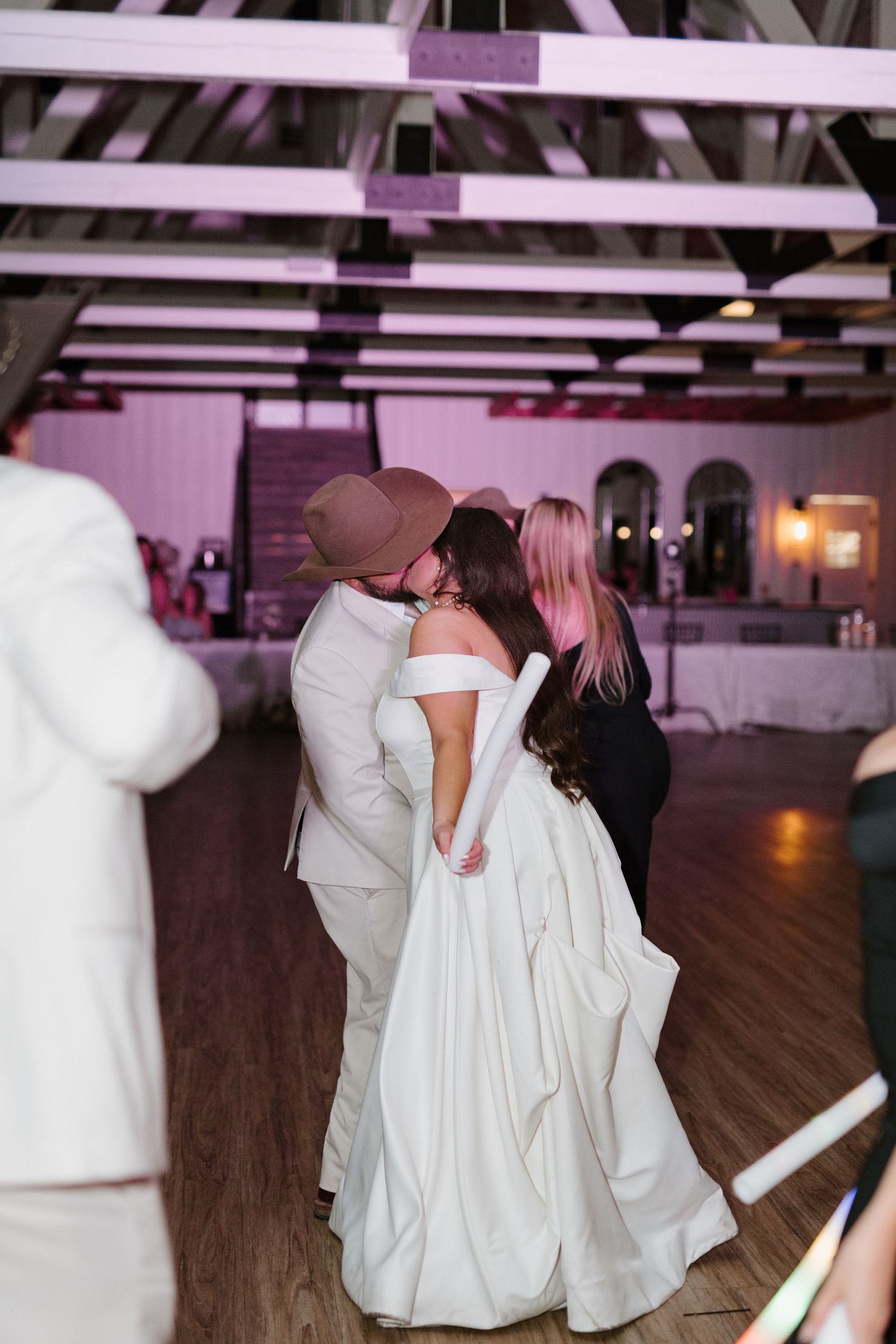 A couple sharing their first dance at a wedding reception under pink-lit wooden beams.