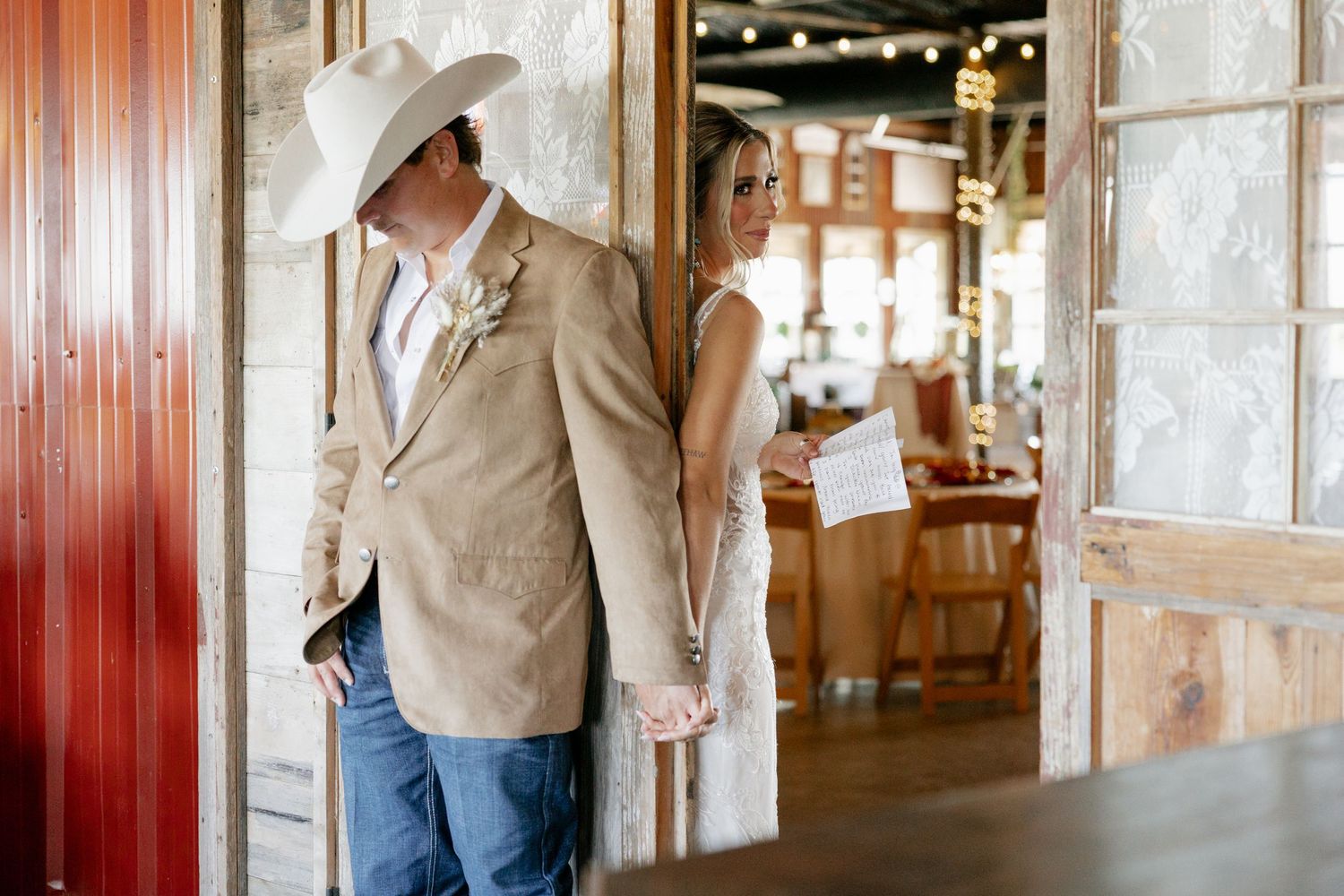 A groom in western attire and cowboy hat stands in a rustic barn doorway.