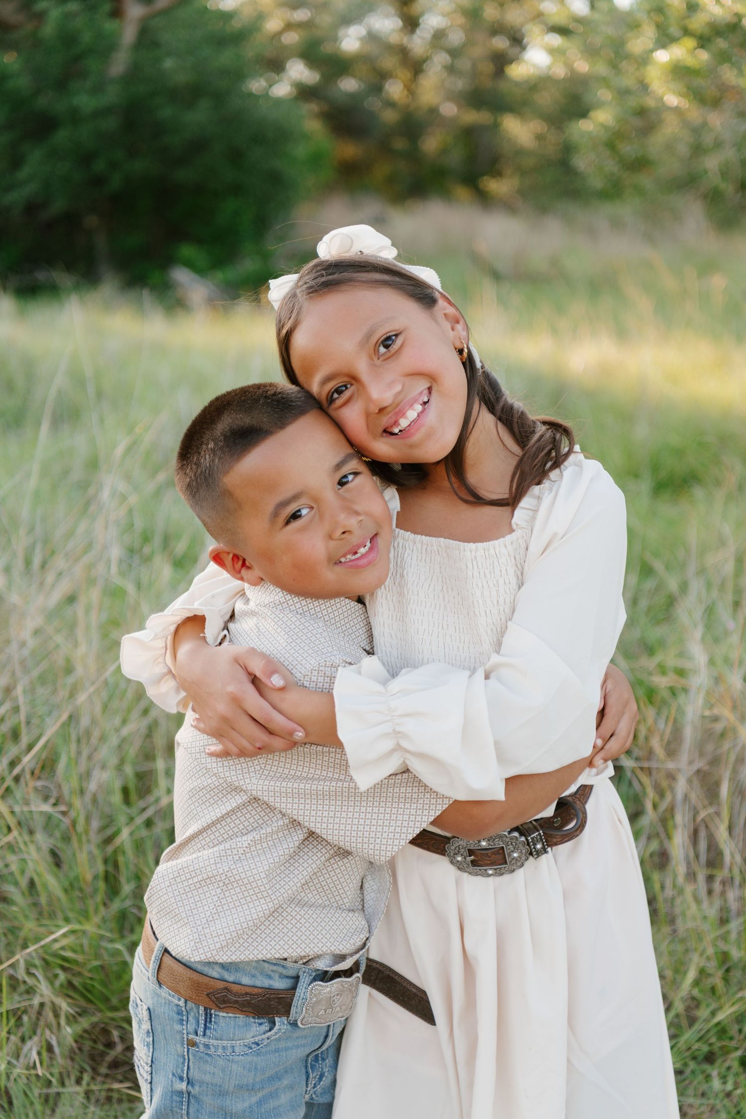 Two siblings embrace and smile warmly while posing together in a golden field at sunset.