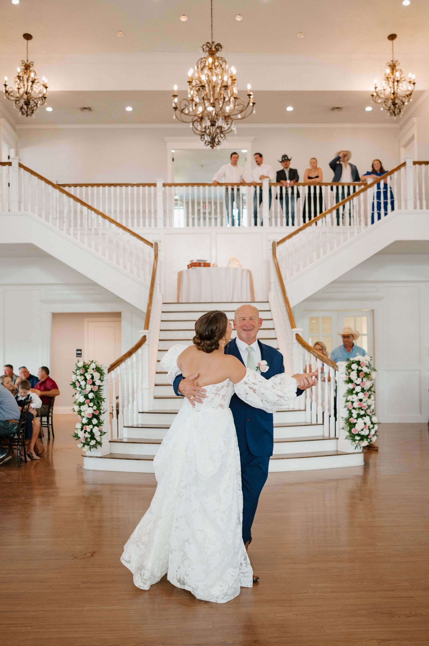 Couple performing first dance in elegant white ballroom with chandelier and staircase.