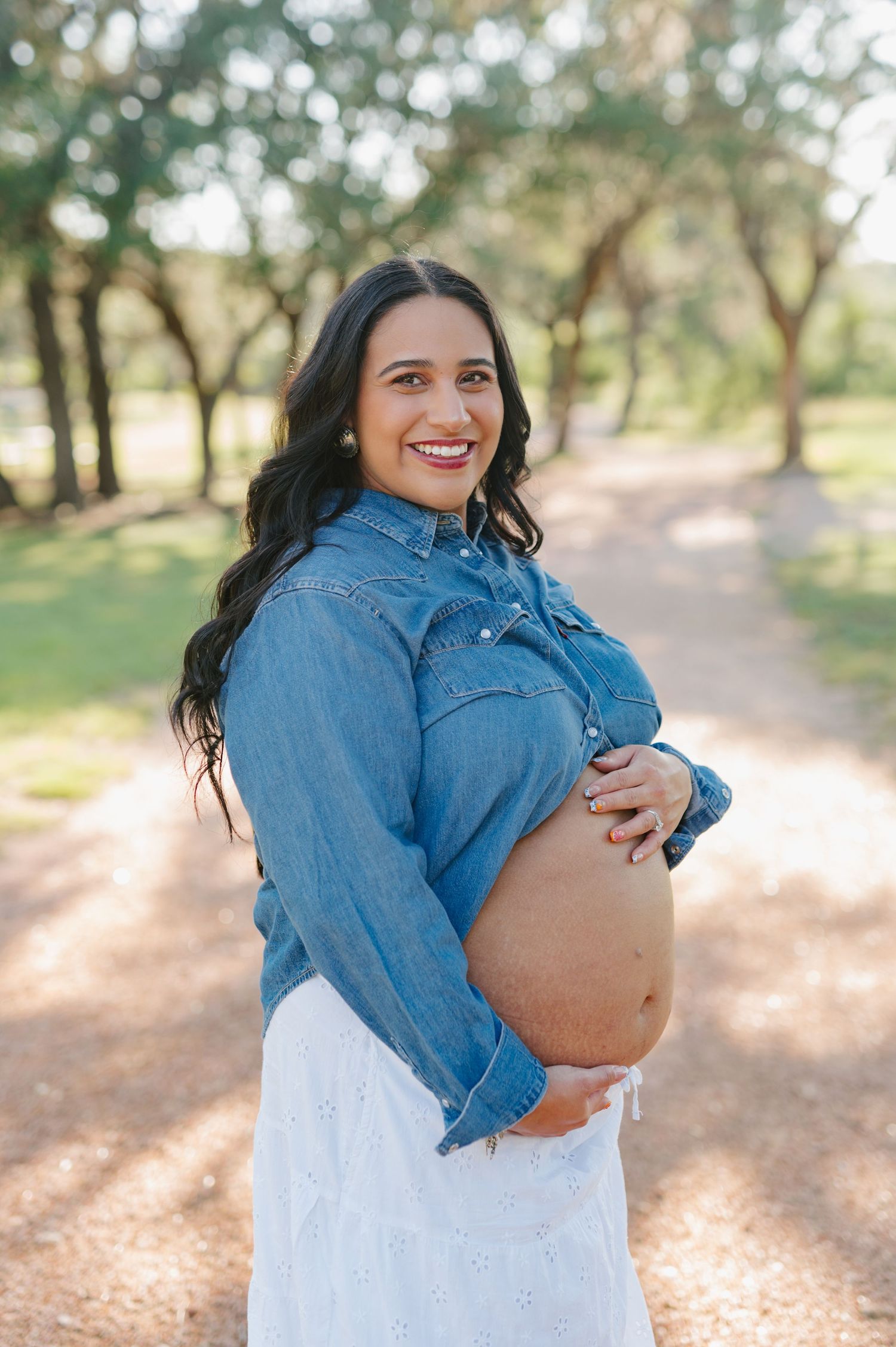 A maternity photoshoot in a park with a denim jacket and white dress against a sunlit wooded backdrop.