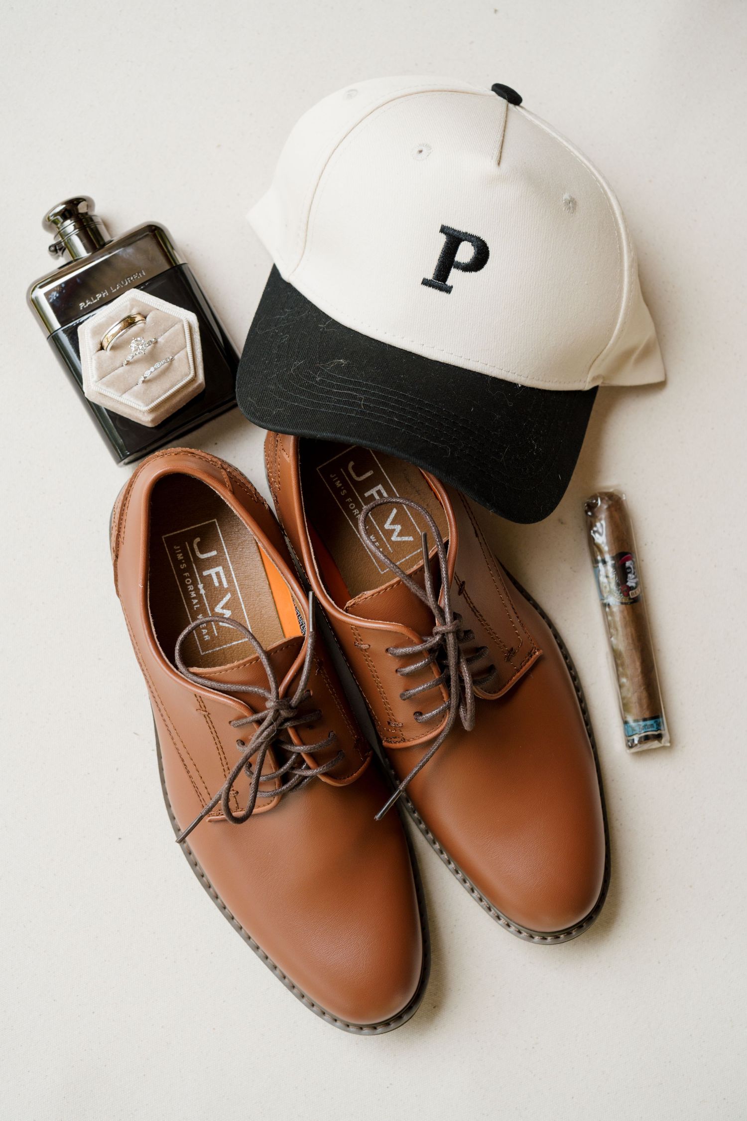 Brown leather dress shoes, white P cap, cologne, ring box, and cigar arranged as groom wedding day accessories.