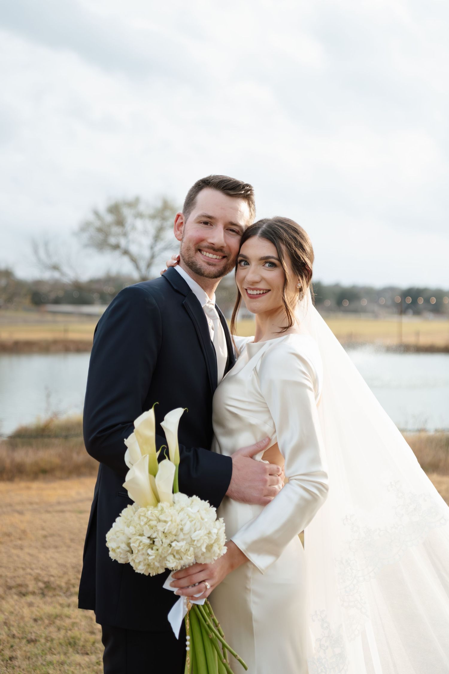 A romantic winter wedding photo series showing a couple in formal attire by a lake with white flowers.