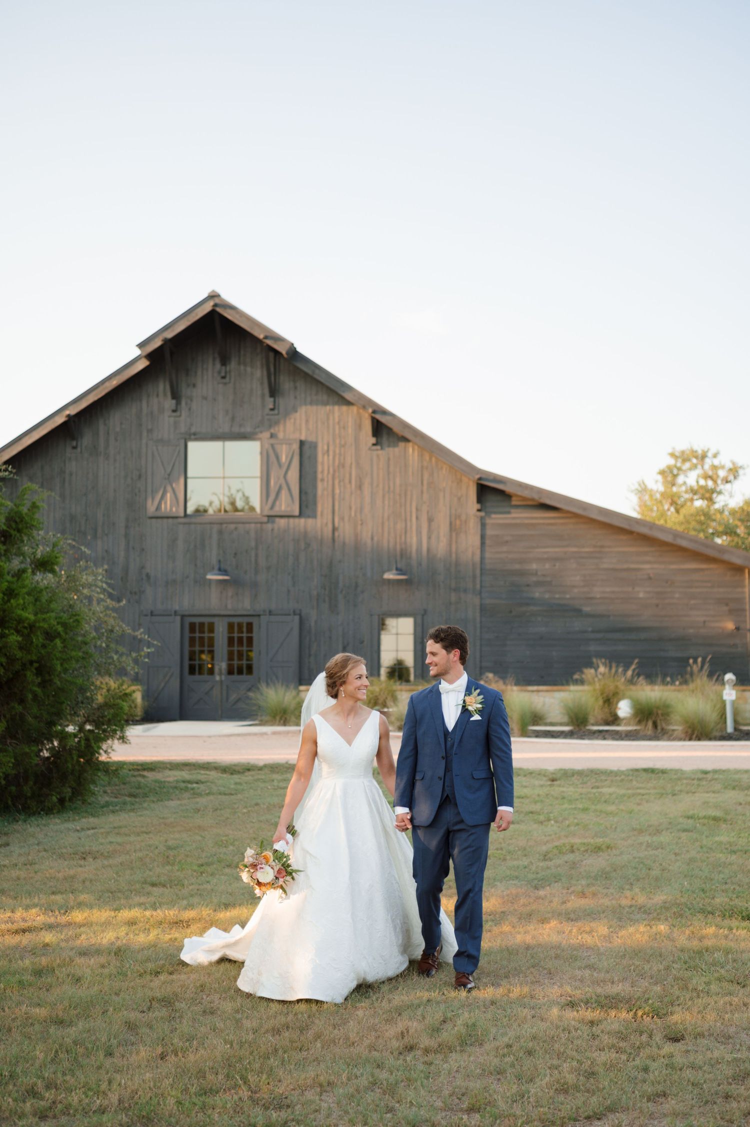 A couple walks together on grass in front of a rustic barn during golden hour wedding photography.