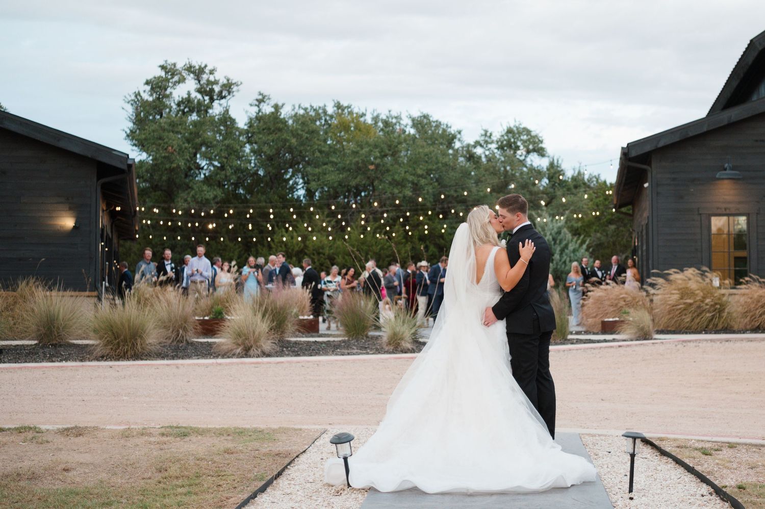 A bride and groom share a romantic embrace in a rustic outdoor setting with string lights and trees in the background.