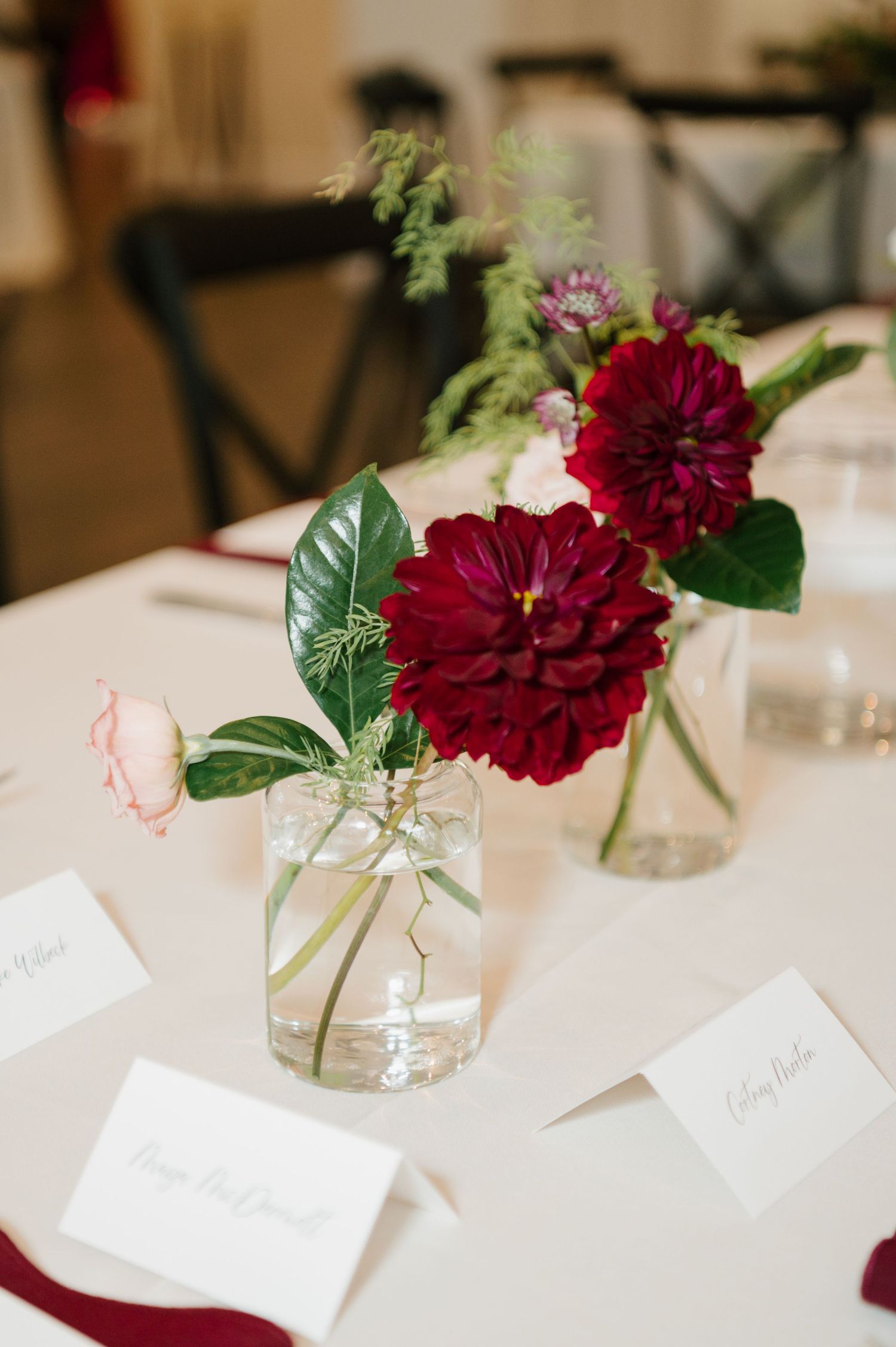 Deep burgundy dahlias in clear glass bud vases with name cards create an elegant table setting at a wedding reception.
