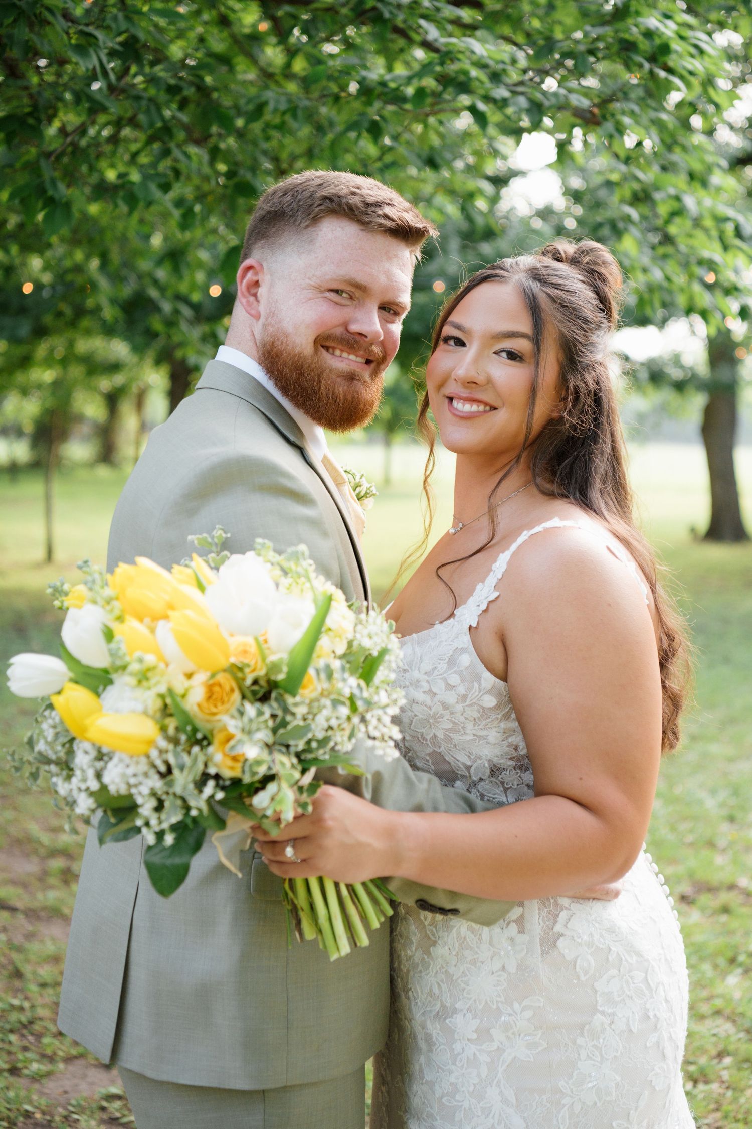 Smiling bride and groom pose together outdoors, bride holding yellow tulip and white flower bouquet, groom in gray suit.