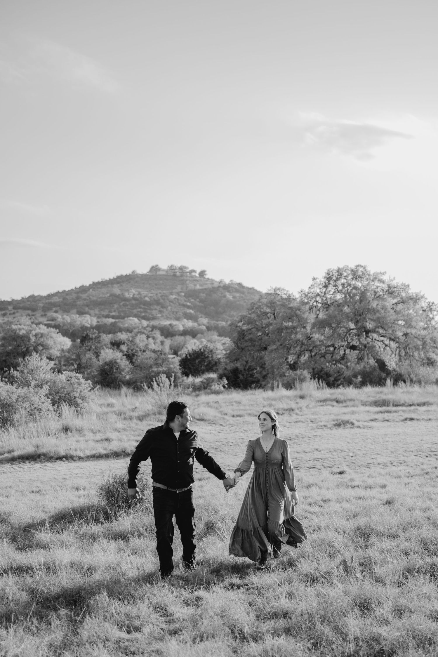 Black and white photo of a couple holding hands walking through a grassy field with hills and trees in the background.