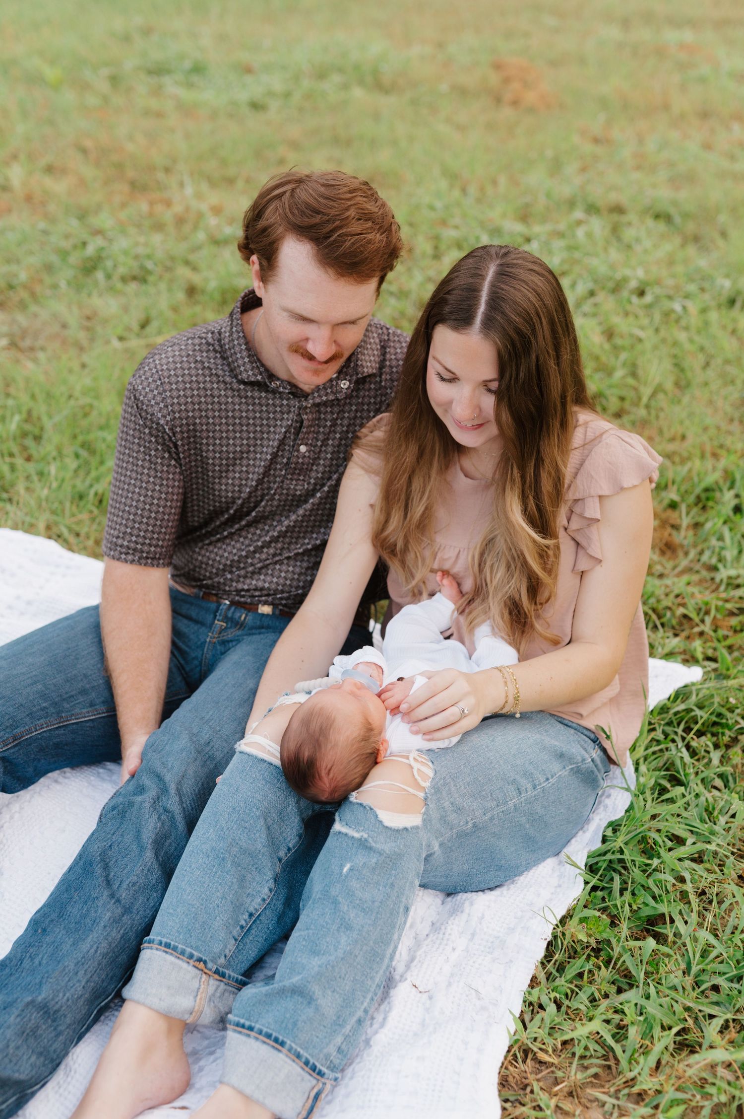 A couple sits together on a white blanket in a grassy field during a casual outdoor photo session.