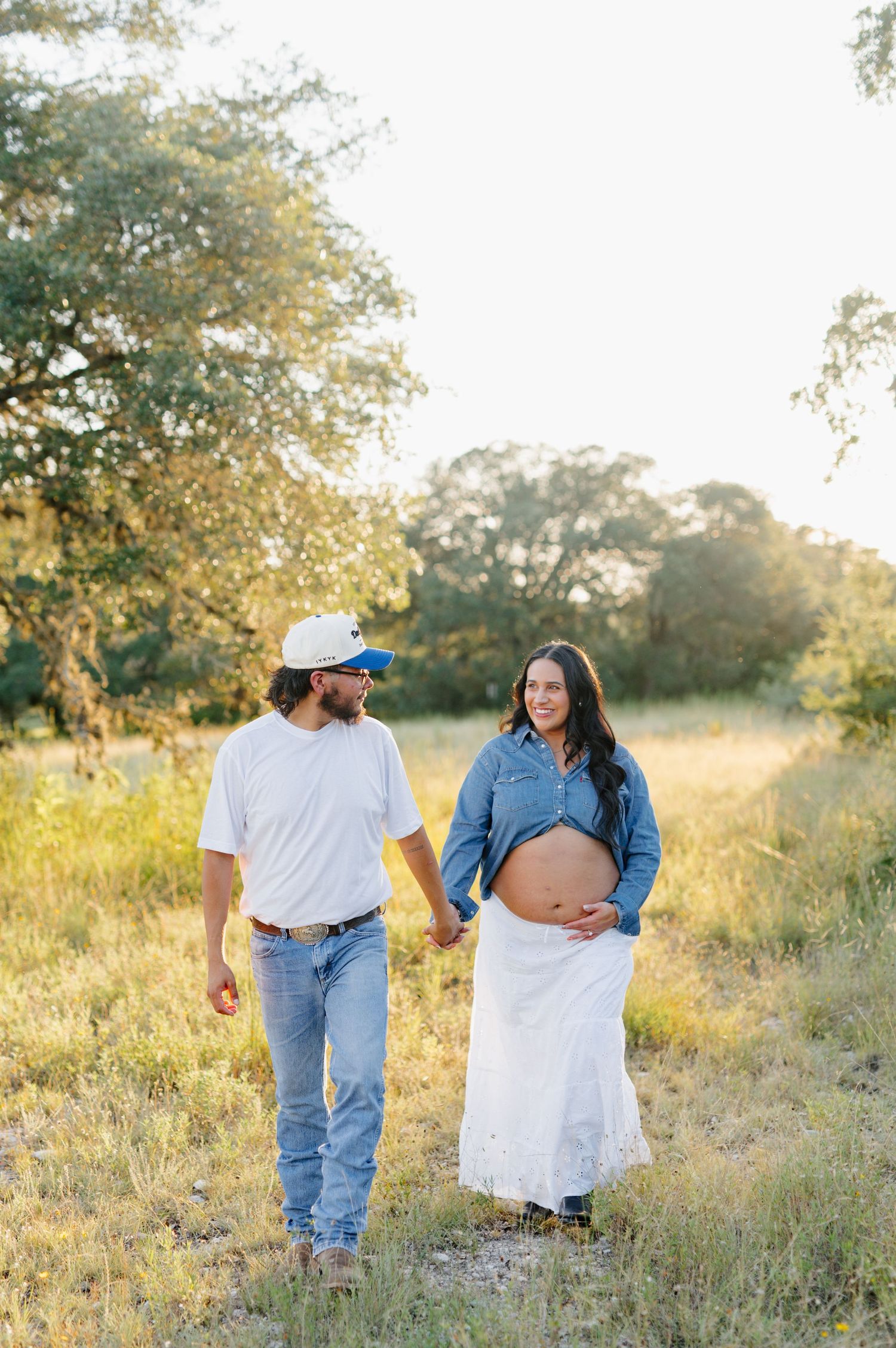 A couple walks through golden grass fields during sunset maternity photoshoot.