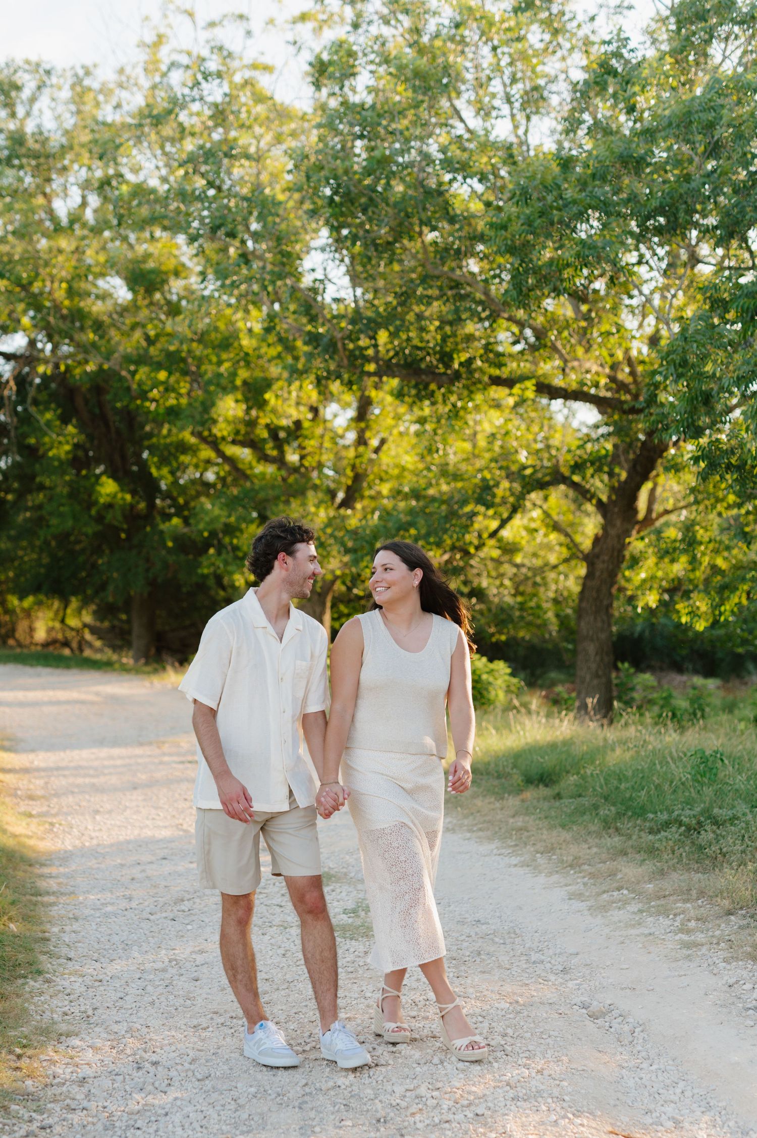 A couple walking hand in hand on a sunlit path surrounded by lush trees.