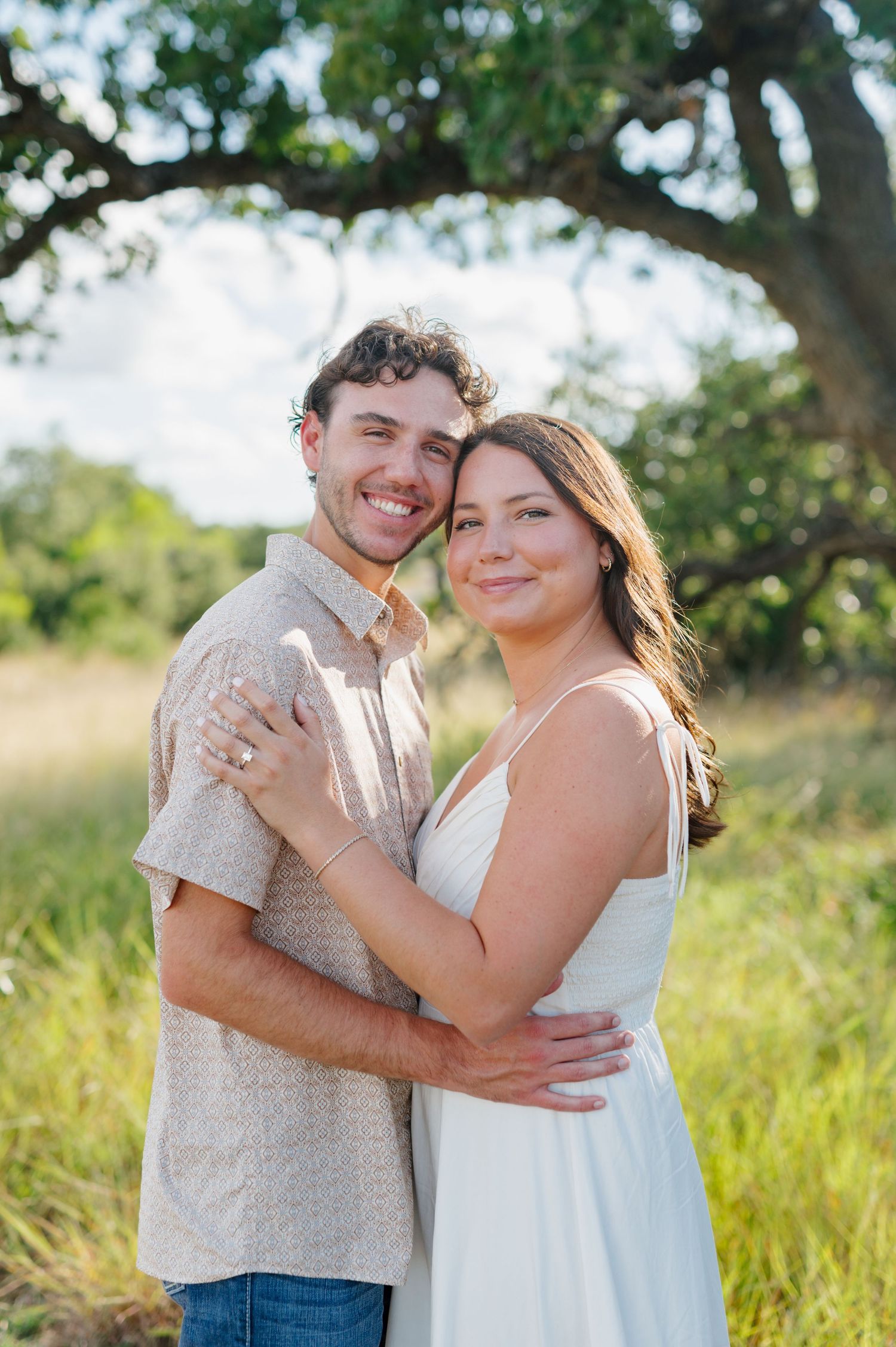 A couple embraces in a sunny field with trees in the background during a romantic outdoor photoshoot.