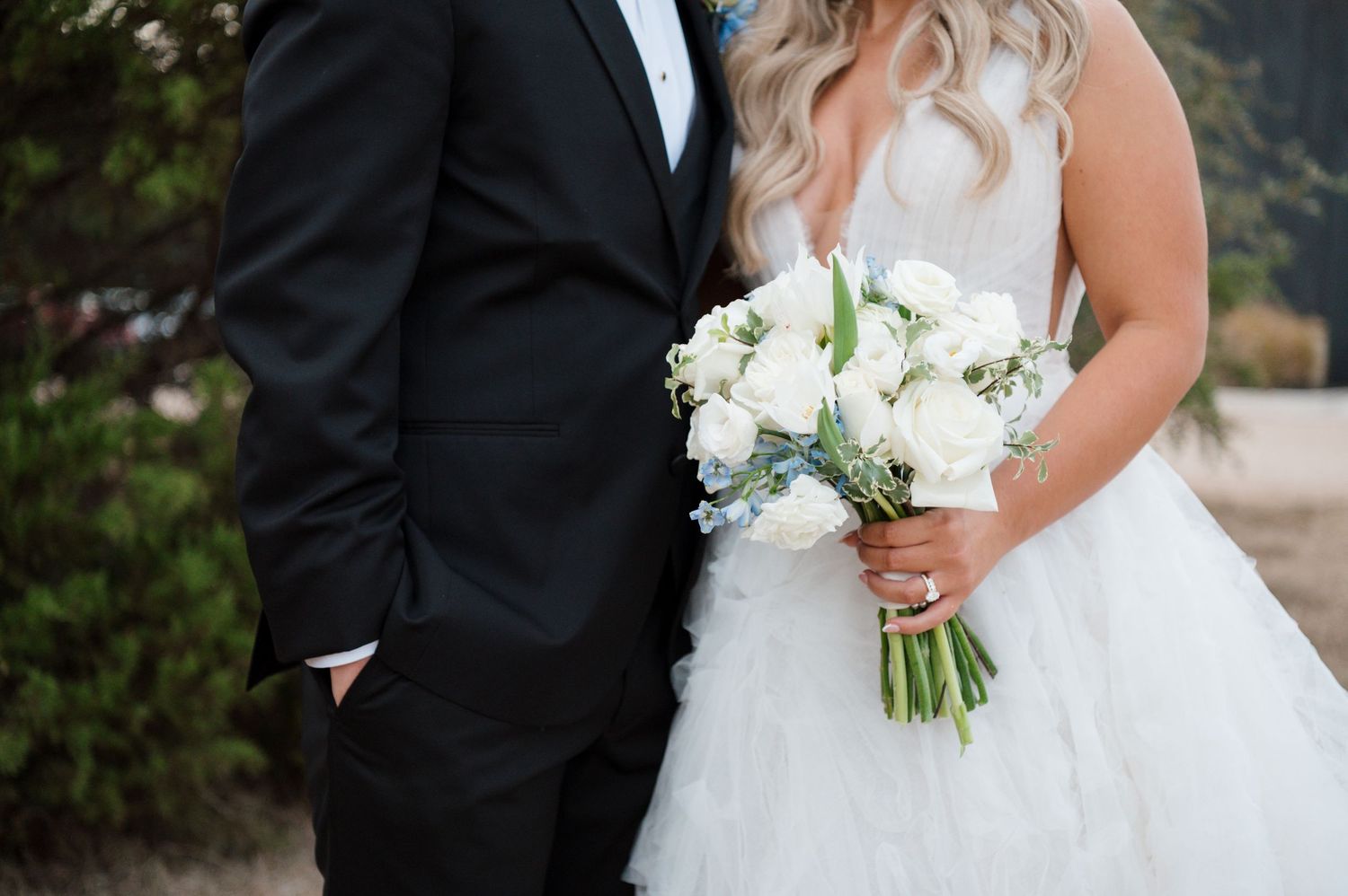 A bride and groom stand together, the bride holding a bouquet of white flowers, in an outdoor setting.