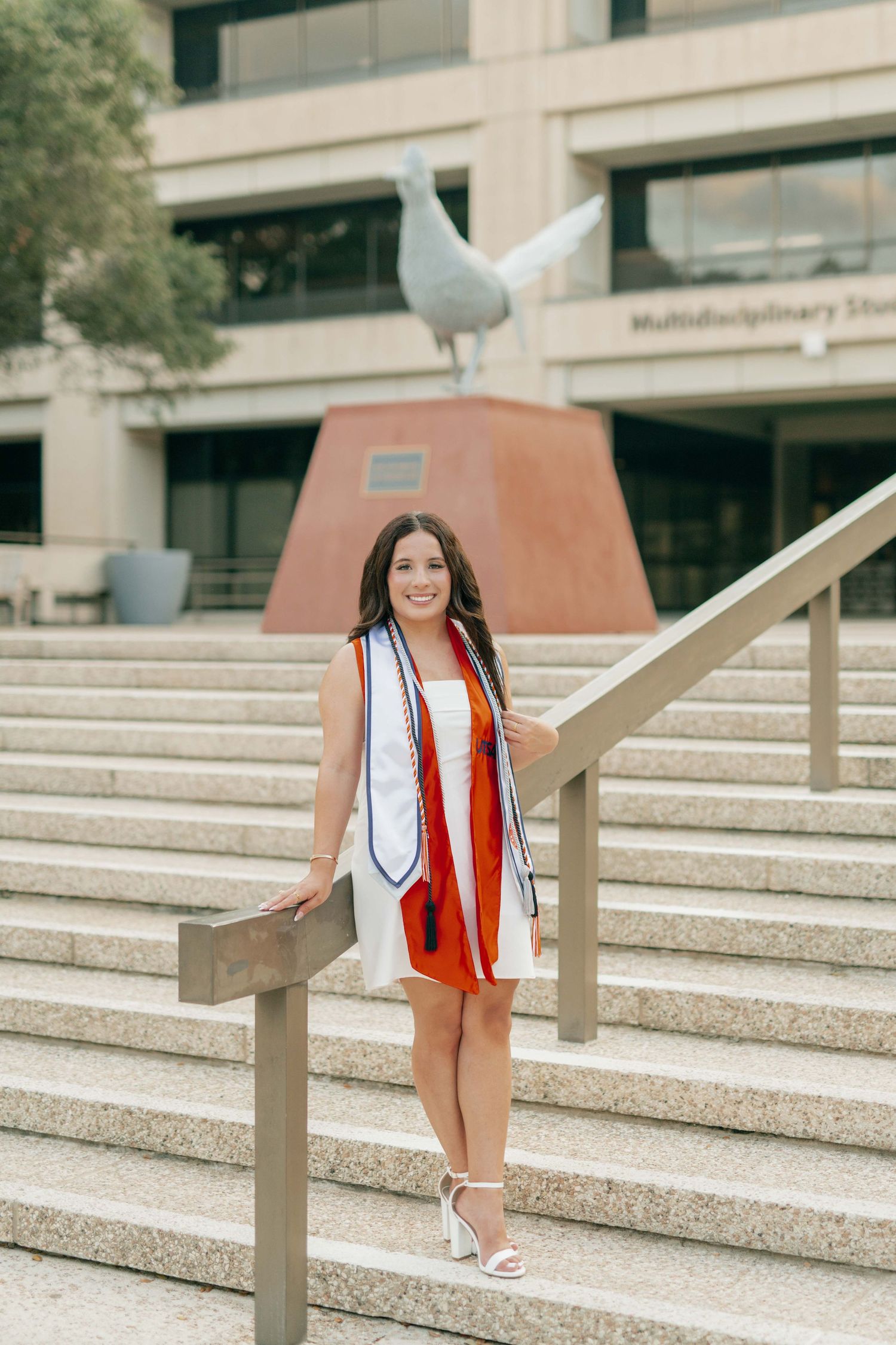 Graduate in white dress and red stole poses on campus steps with seagull statue.