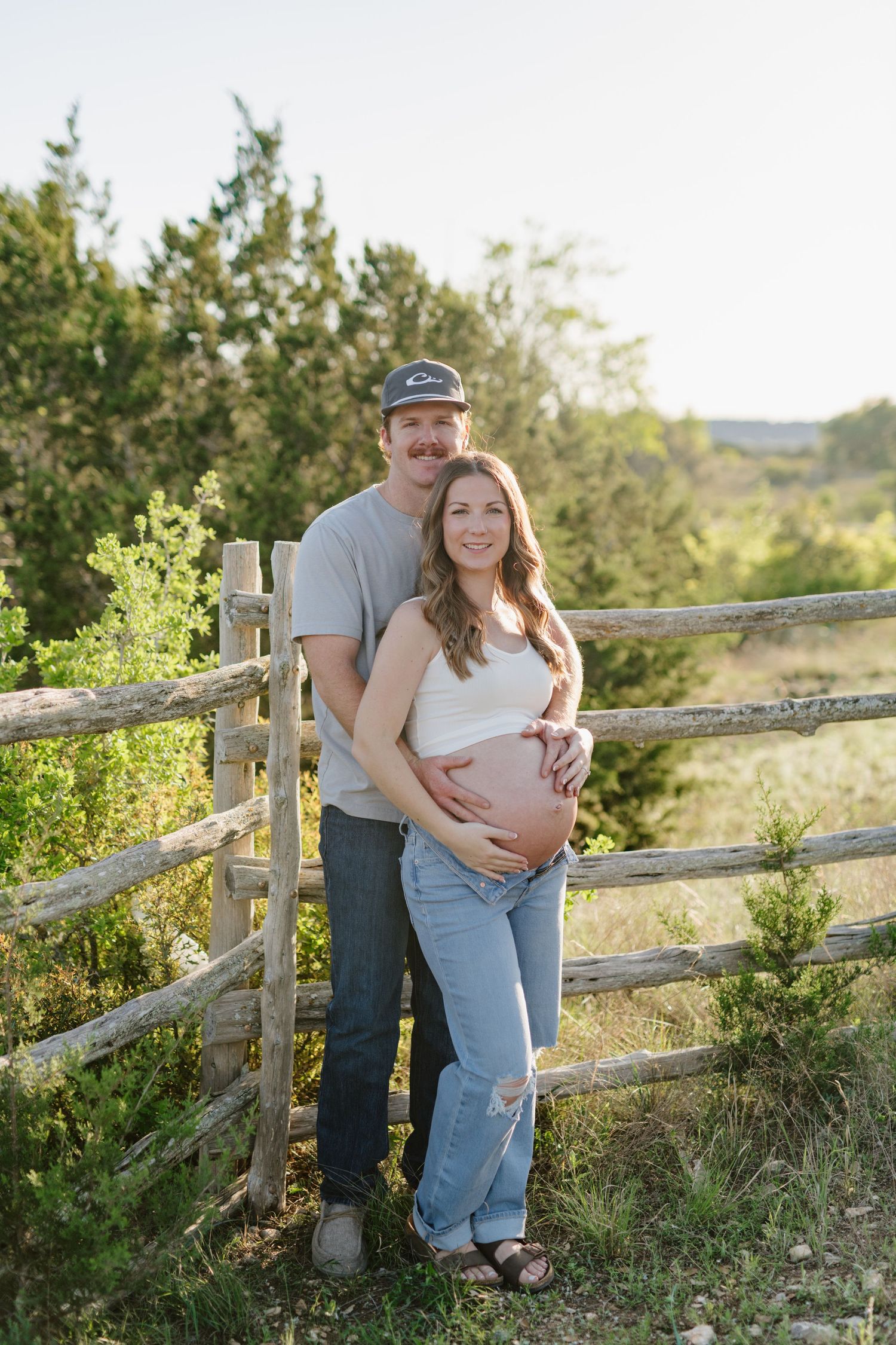 Expectant couple poses by rustic wooden fence during outdoor maternity photography session in sunlit field.