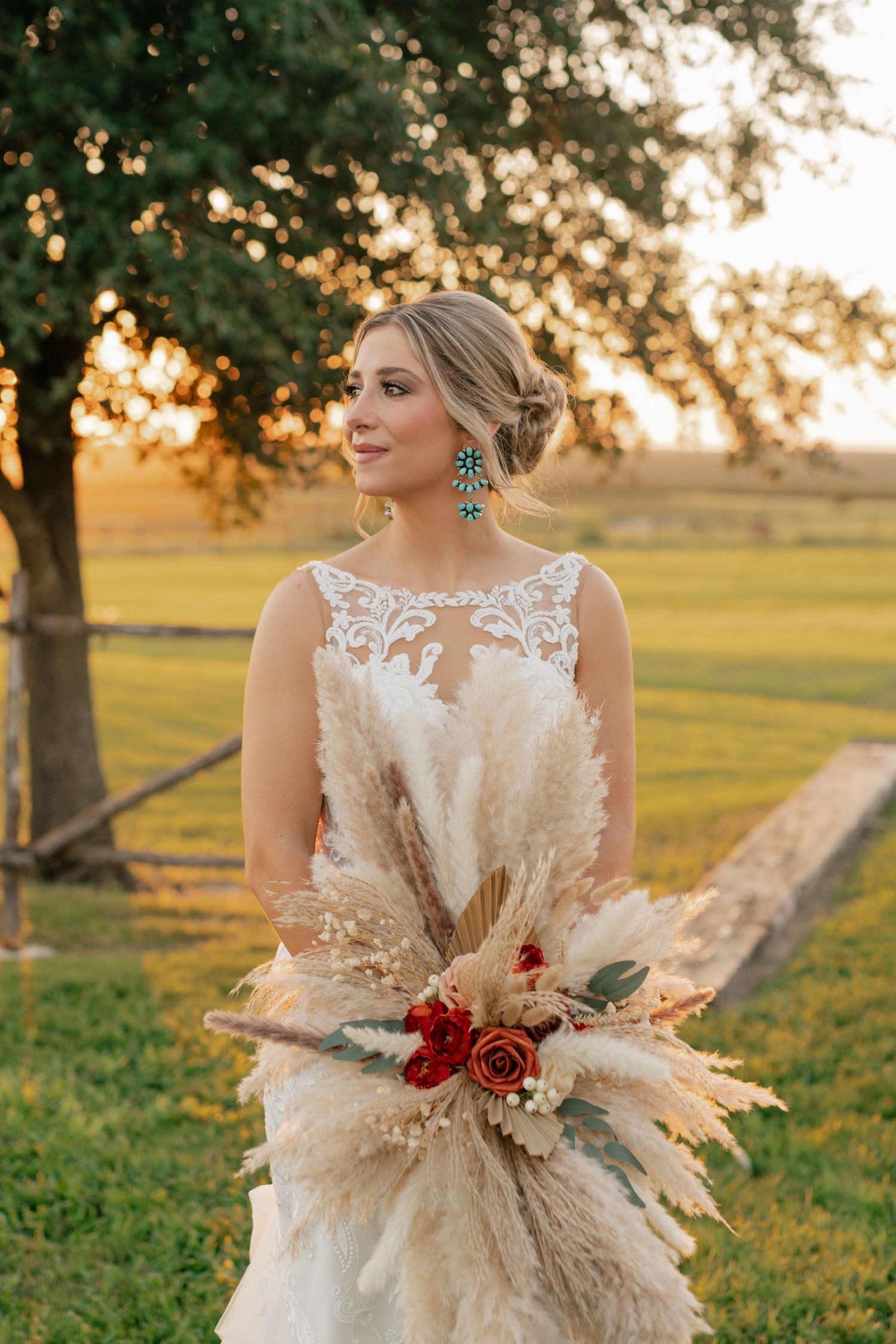 Bride in lace wedding dress holds elegant bouquet with pampas grass and roses during sunset in outdoor setting.