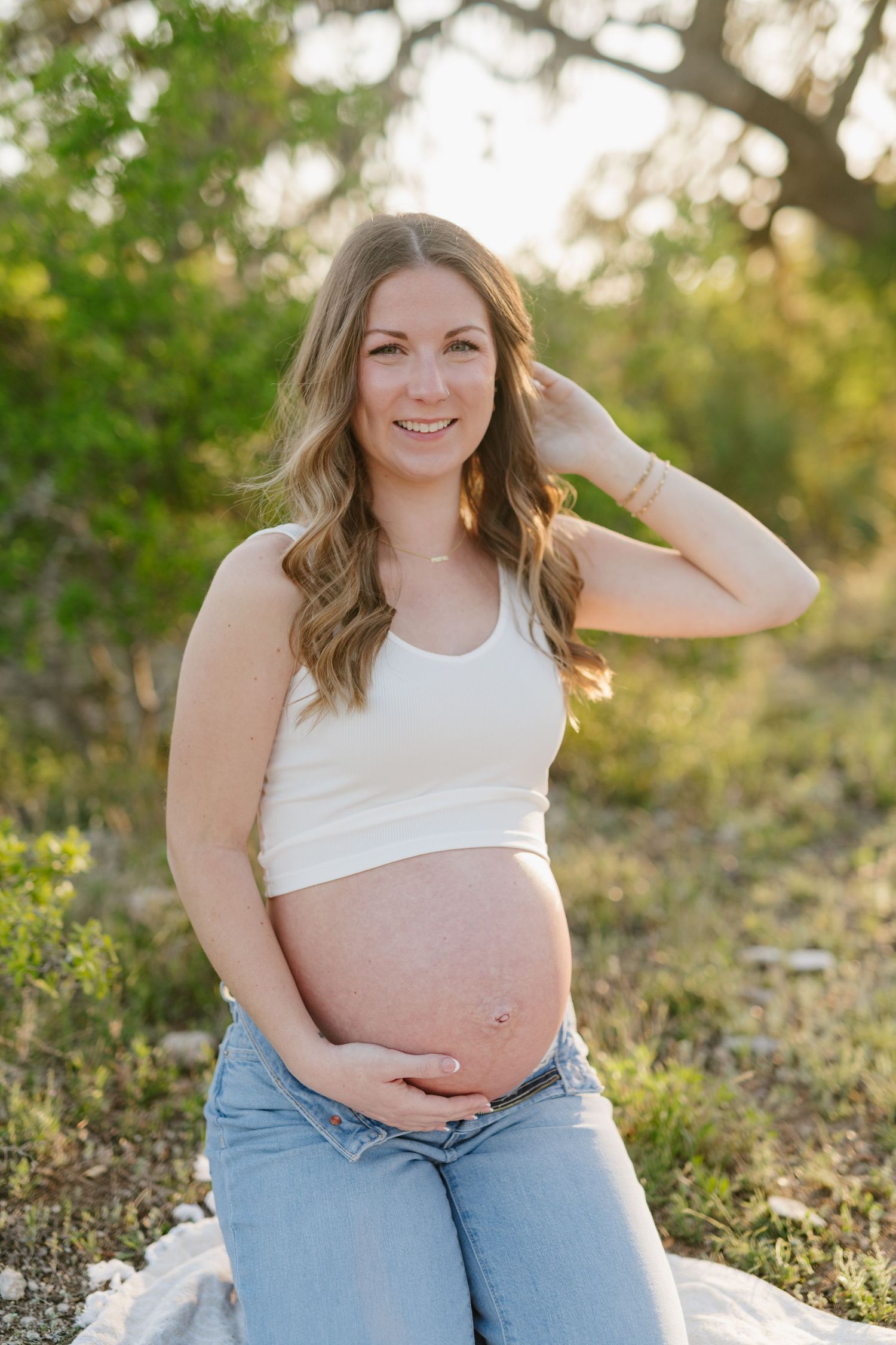 Pregnant woman in casual attire sits outdoors during golden hour while holding her baby bump.