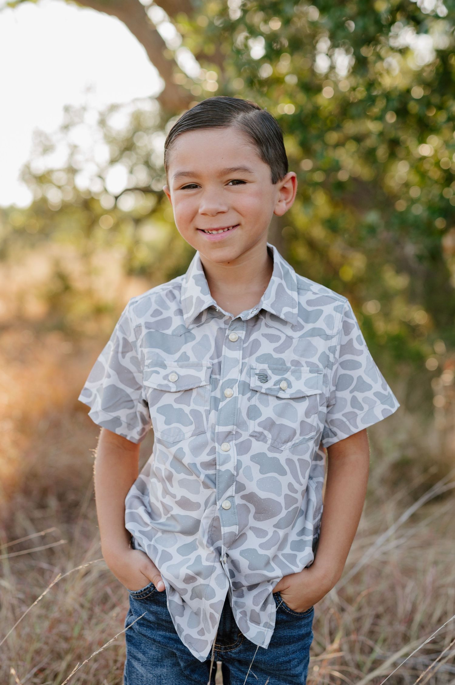 A child wearing a gray patterned button-down shirt and jeans poses outdoors against a blurred natural background.