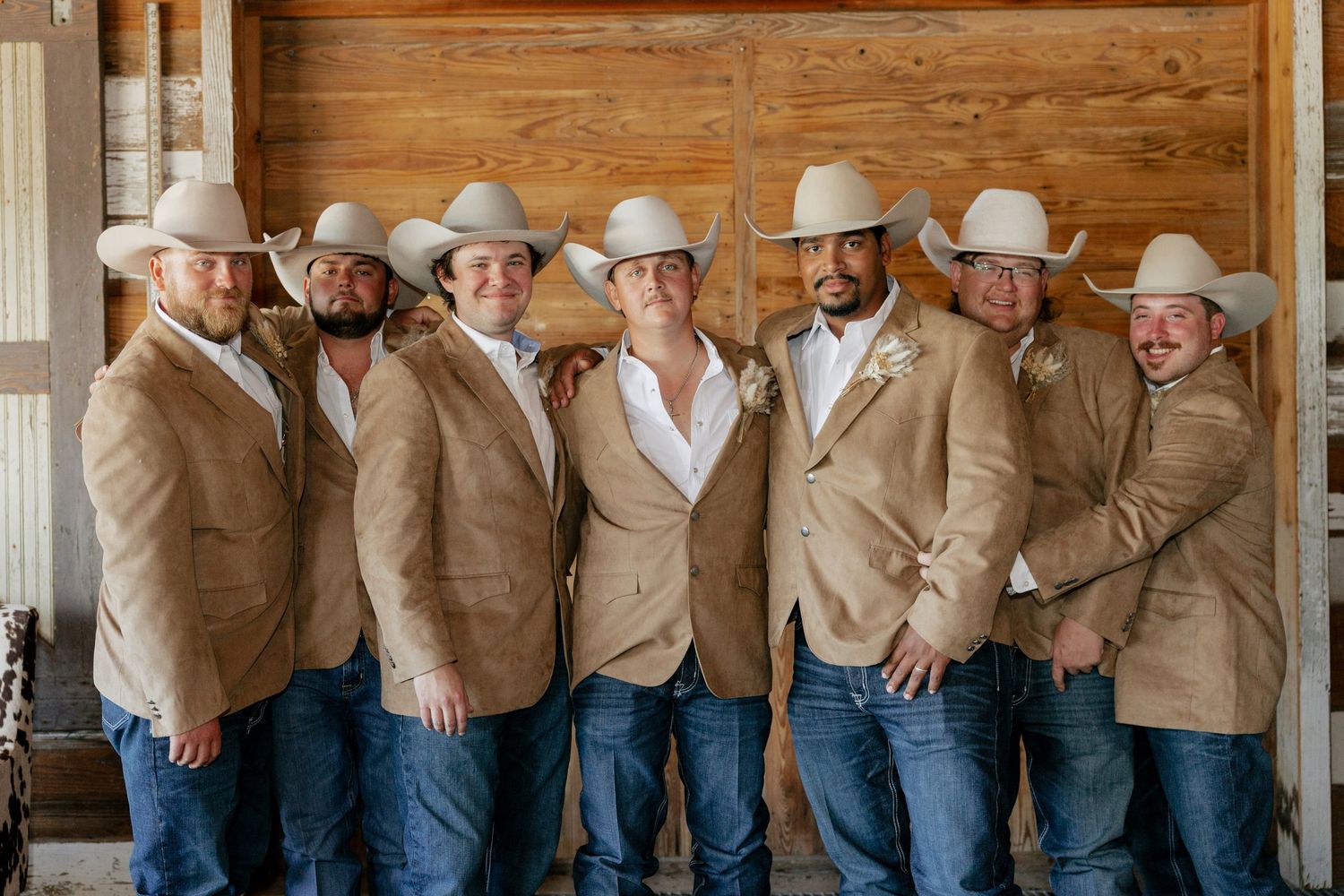 Groomsmen in coordinated tan jackets and cowboy hats pose together at a rustic venue.