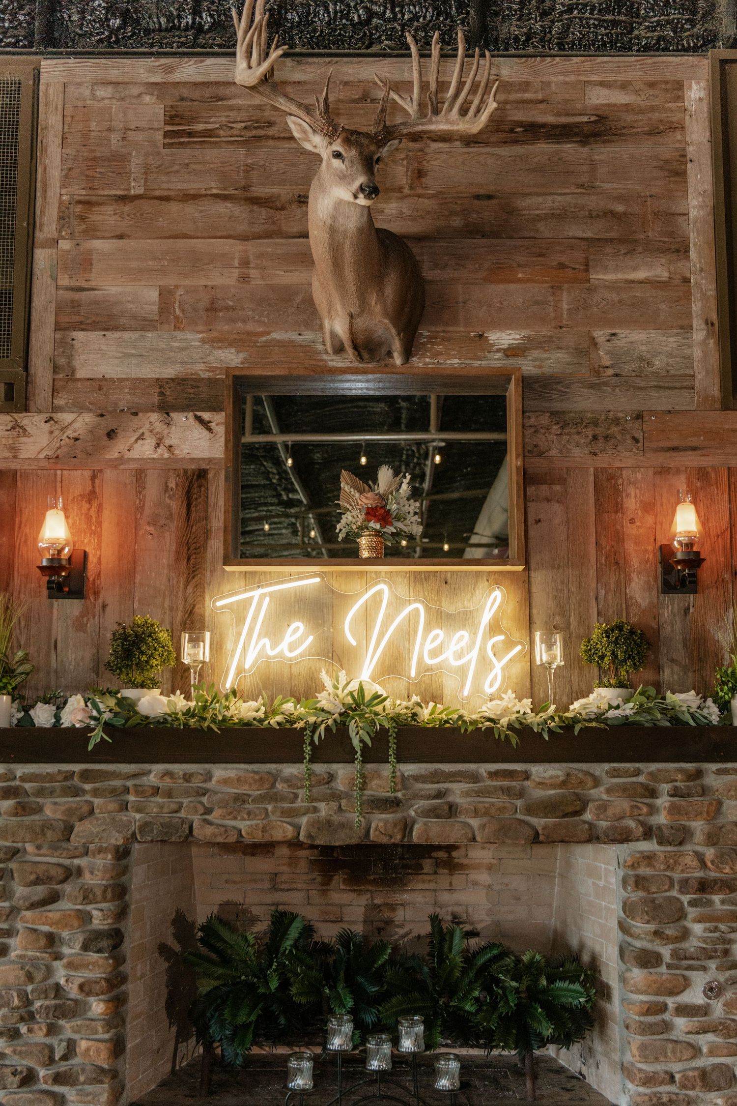 Rustic wedding decor featuring mounted deer head, neon sign, and stone fireplace with greenery and candle accents.