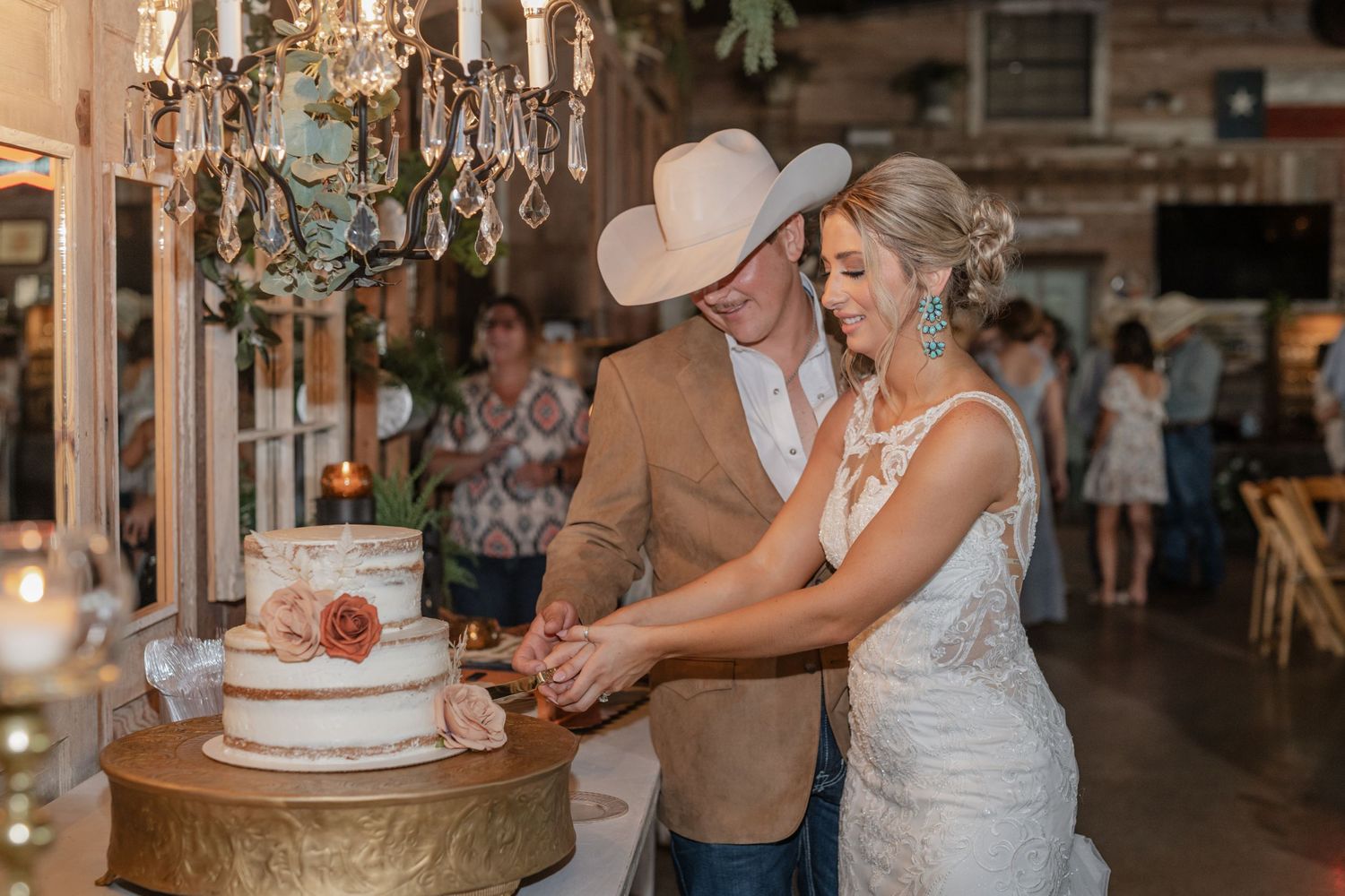 Newlyweds cutting their elegant white wedding cake under vintage chandelier in rustic venue.
