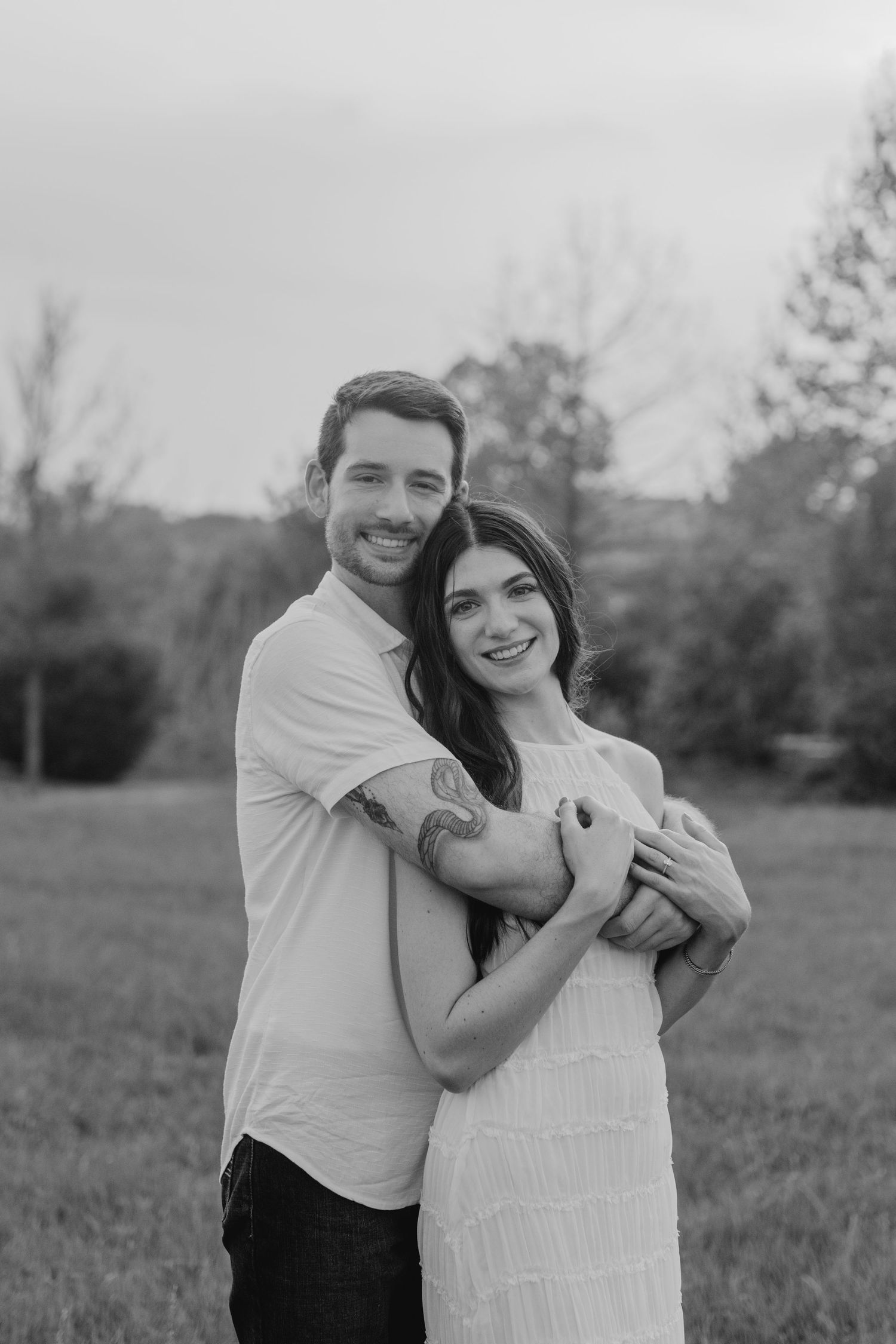 A romantic black and white portrait of a couple embracing in a field with trees behind them.