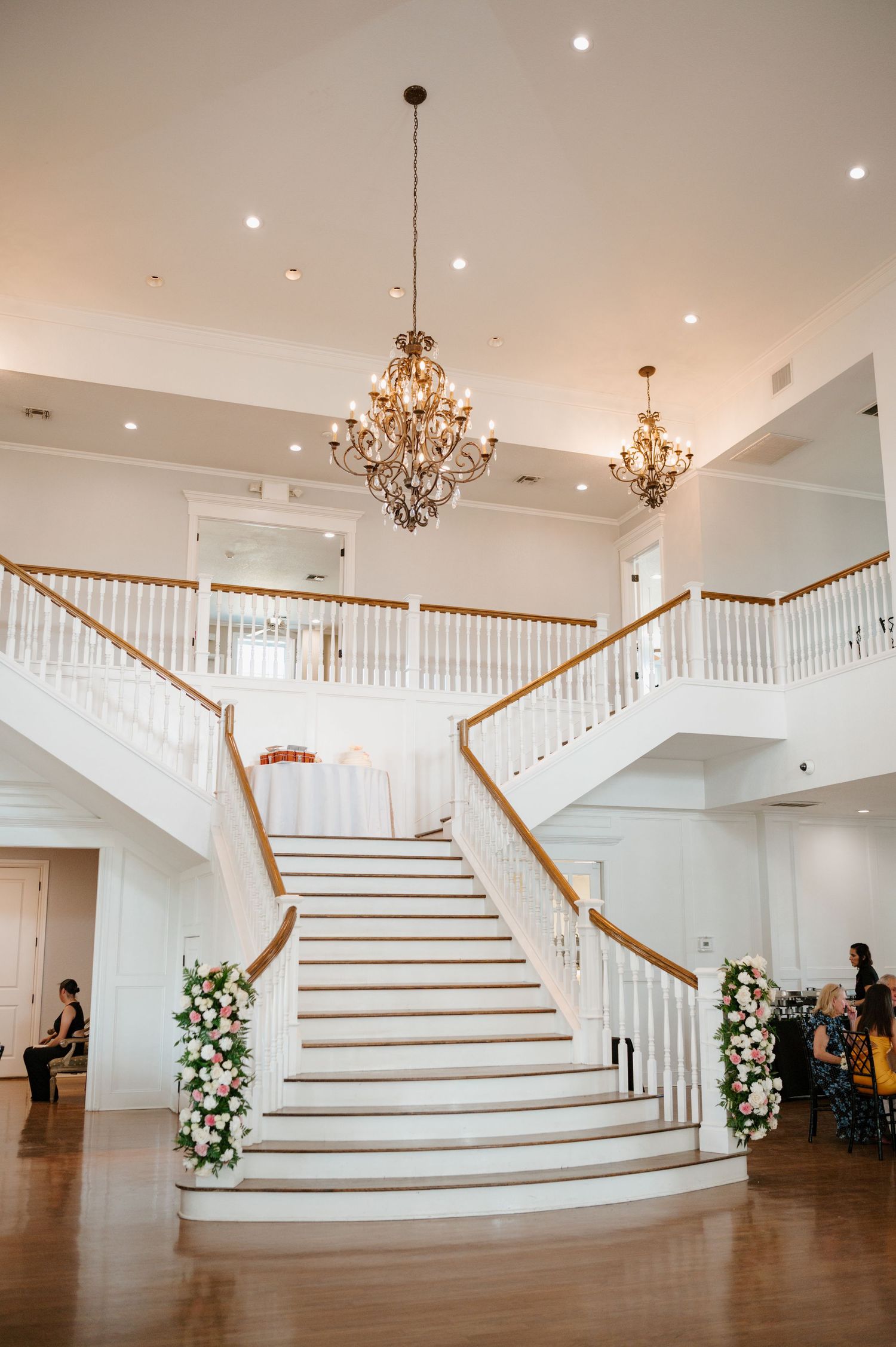 Elegant grand staircase with white railings and wooden accents adorned with floral arrangements and crystal chandeliers.