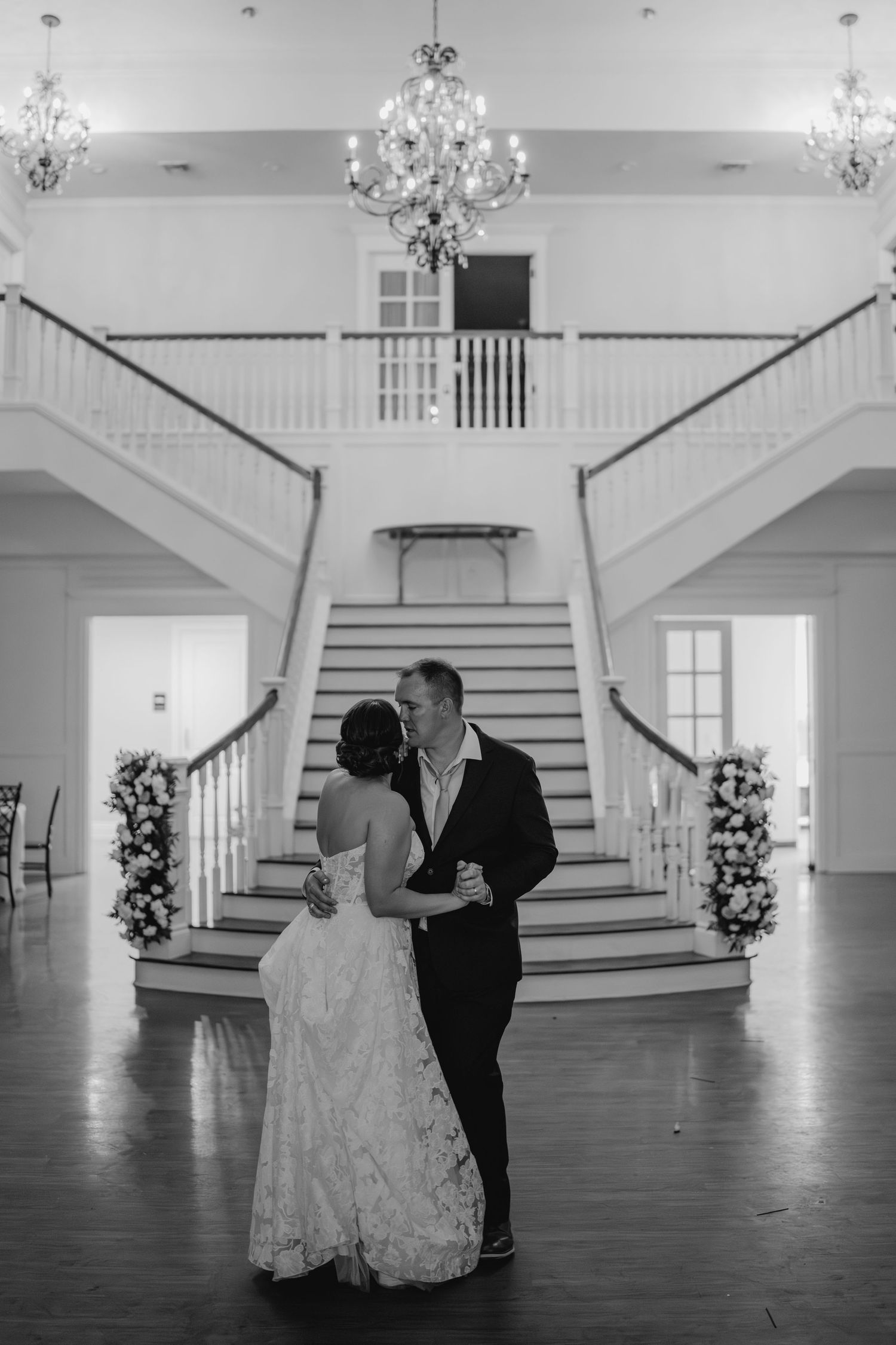 Black and white photo of couple dancing in elegant venue with grand staircase.
