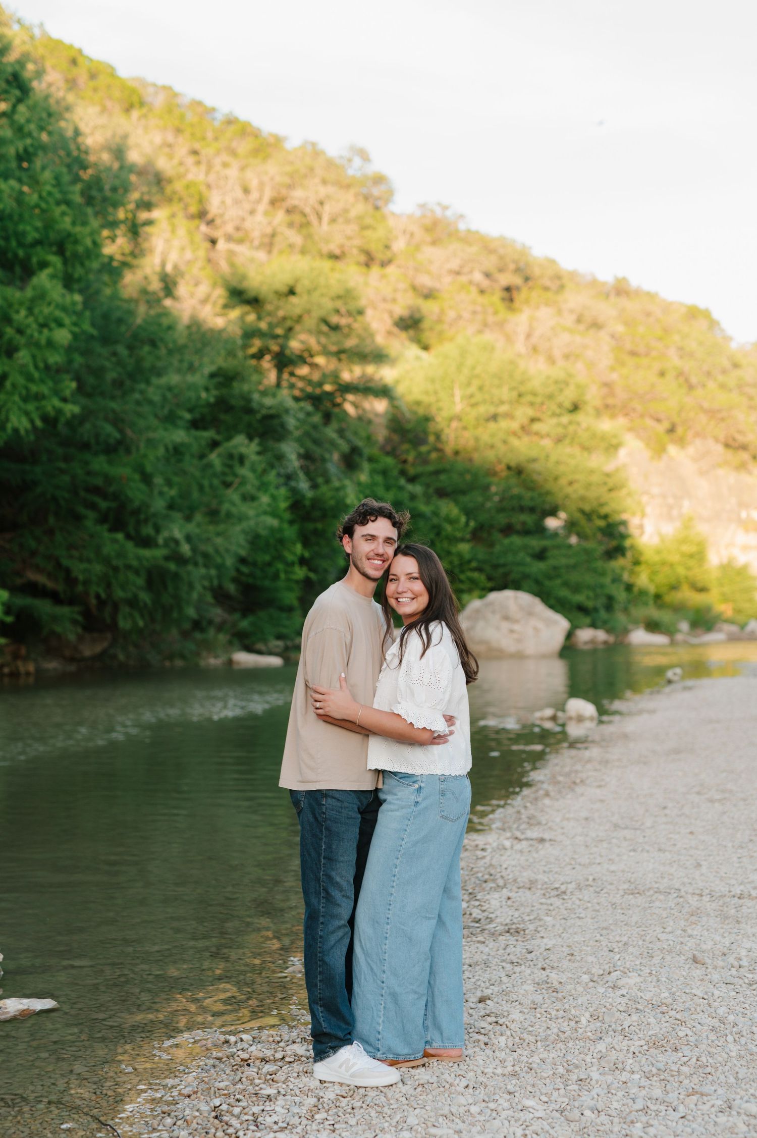 A couple stands together on a rocky riverbank during golden hour with tree-covered hills in the background.